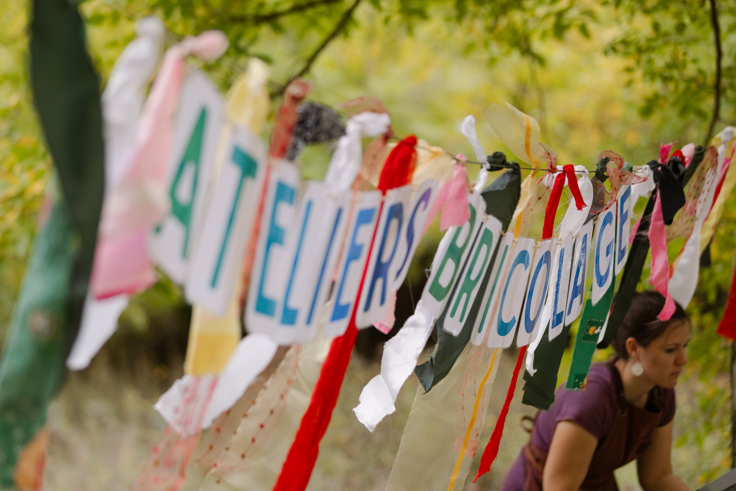 Bandes de tissu coloré et lettres formant le mot 'ATELIERS BRICOLAGES' suspendues entre deux arbres dans une forêt.