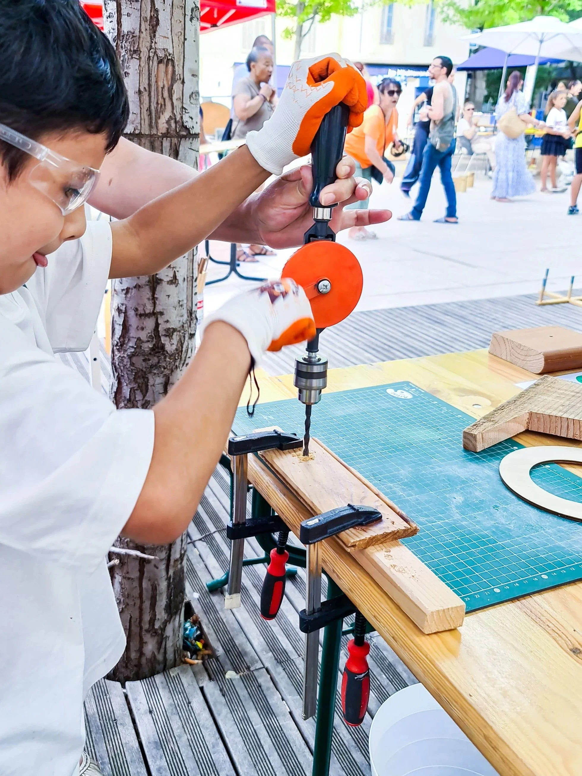 Un enfant utilise une perceuse manuelle pour percer un morceau de bois lors d'une activité extérieure, avec plusieurs personnes en arrière-plan.