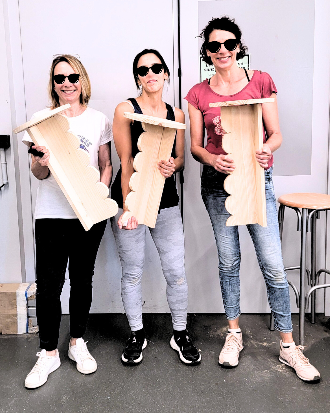 Trois femmes debout, portant des lunettes de soleil, tenant des tables en bois fabriquées par elles-mêmes dans un atelier de menuiserie