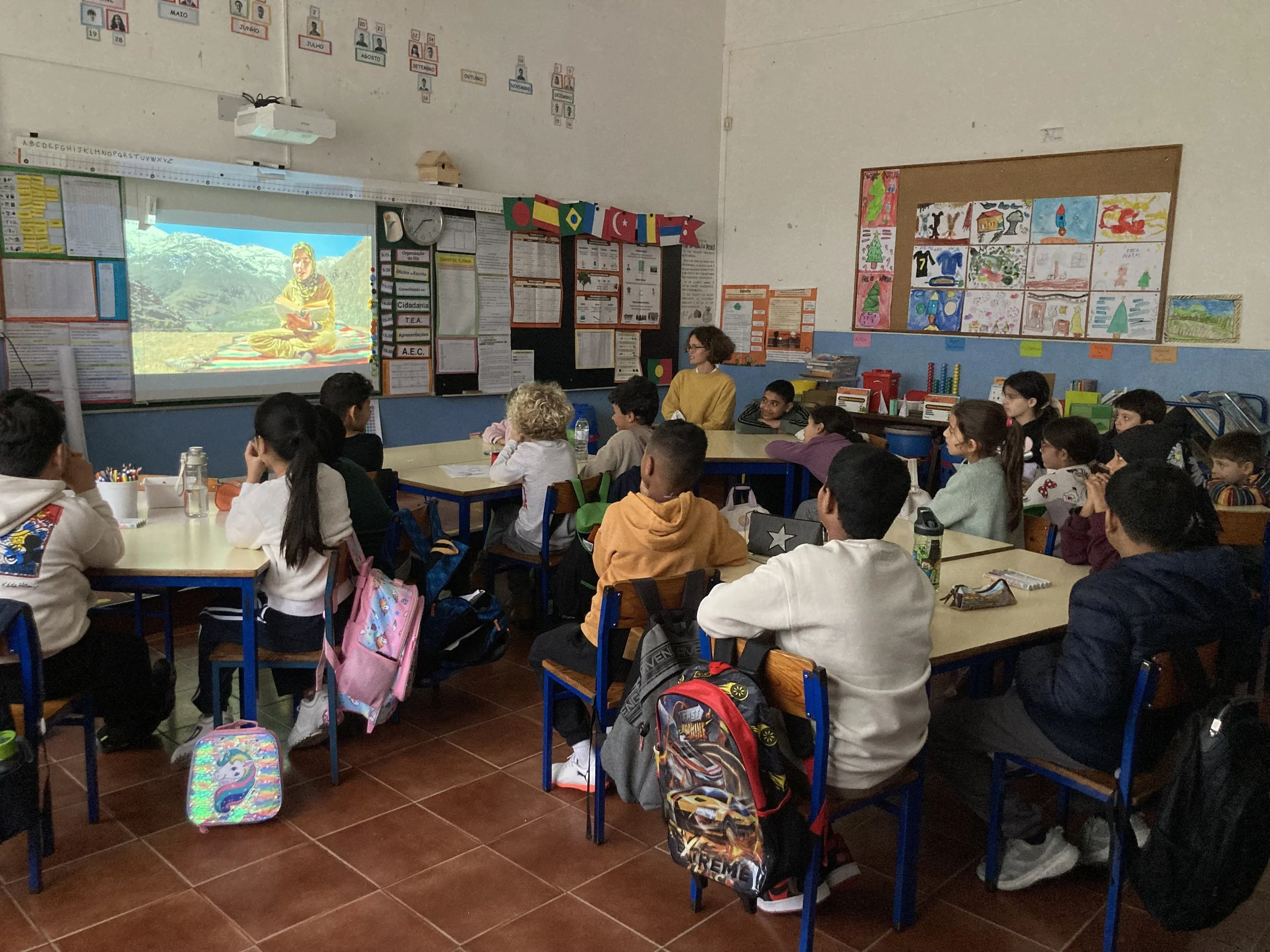 Um grupo de crianças sentadas na sala de aula, a ver um filme, projetado num quadro branco. As crianças estão sentadas em cadeiras de madeira, com estrutura metálica azul, onde penduraram as mochilas. Estão, em grupos de quatro, à volta de mesas de m