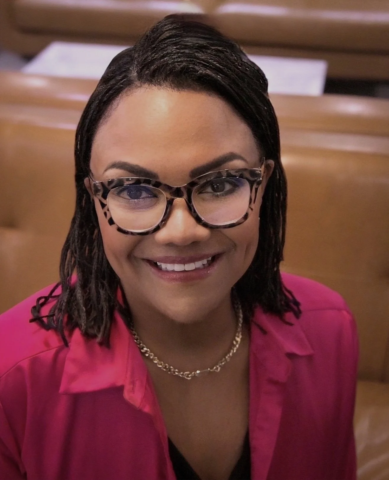 A photo of Michelle Courtney Berry wearing glasses, a pink blazer, a pearl necklace, and a gold chain, smiling in an indoor setting with wooden furniture in the background.