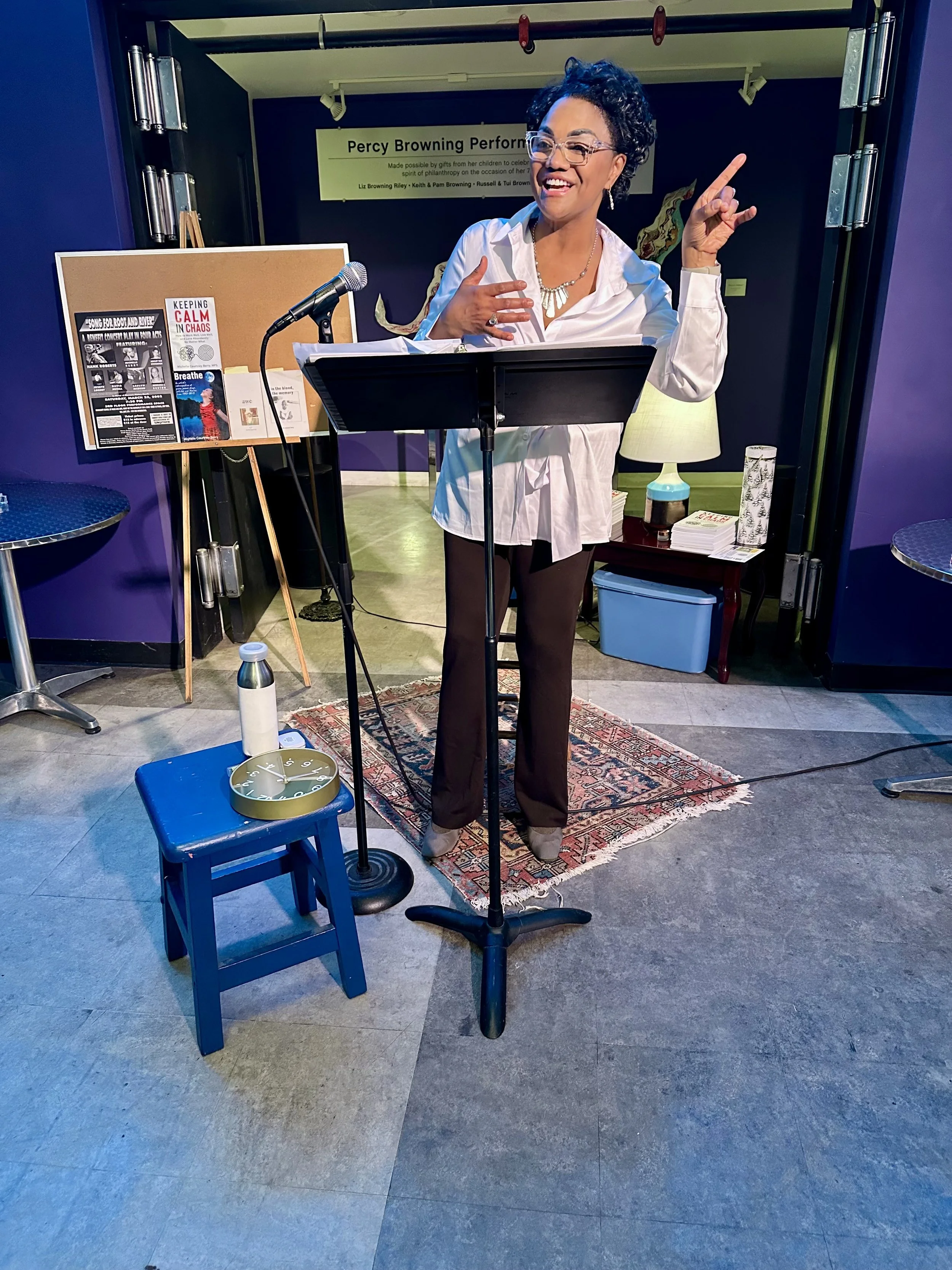 A woman standing at a music stand, smiling and gesturing with her hand while singing or speaking, with a corkboard, a microphone, and various objects around her in a room with purple walls and a sign about a performance in the background.