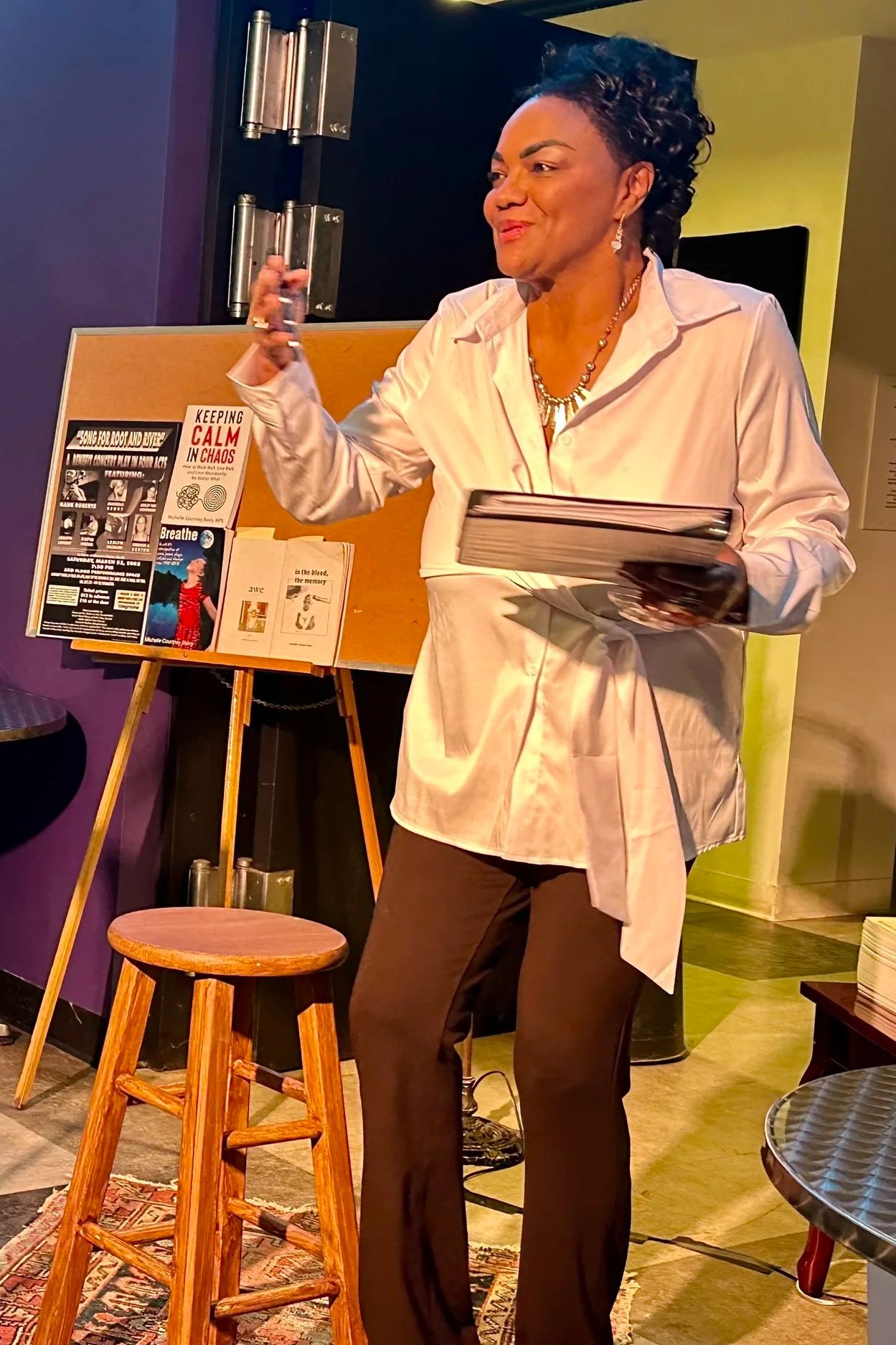 A woman in a white shirt holding a glass of wine and a book, standing in a cozy indoor space with posters on a wooden easel and a small wooden stool nearby.