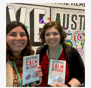 Two women smiling and holding books titled 'Keeping Calm in Chaos' at an event with a colorful, abstract mural in the background.