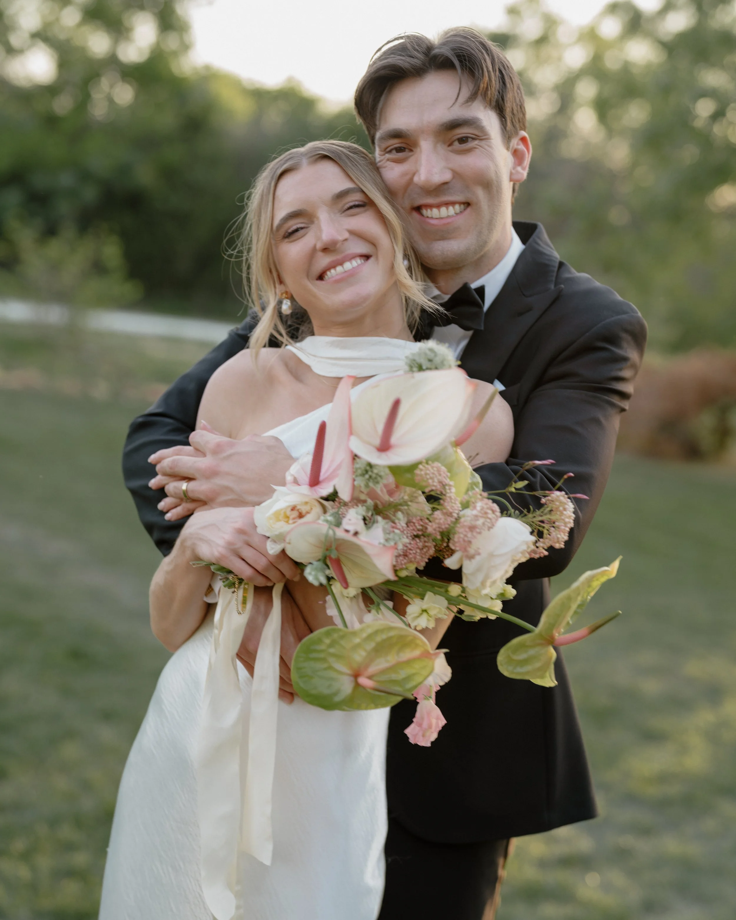 A newlywed couple at Barr Mansion smiling and hugging outdoors during the daytime, with the bride holding a bouquet of pink and white flowers.