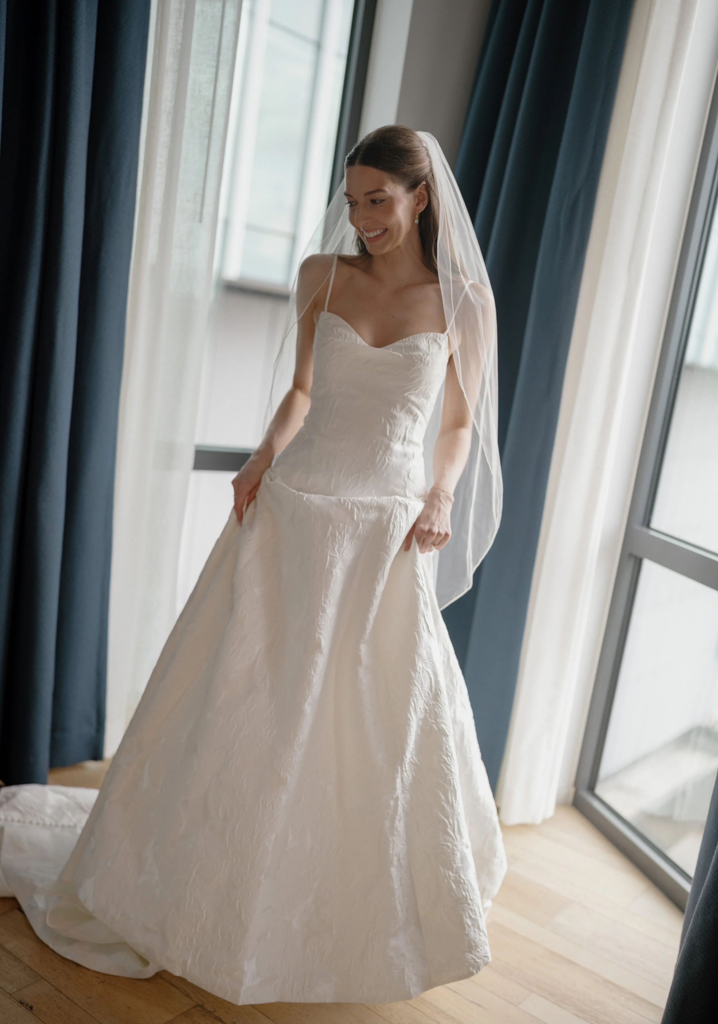 A bride in a white wedding gown with a veil standing by a large window, smiling.