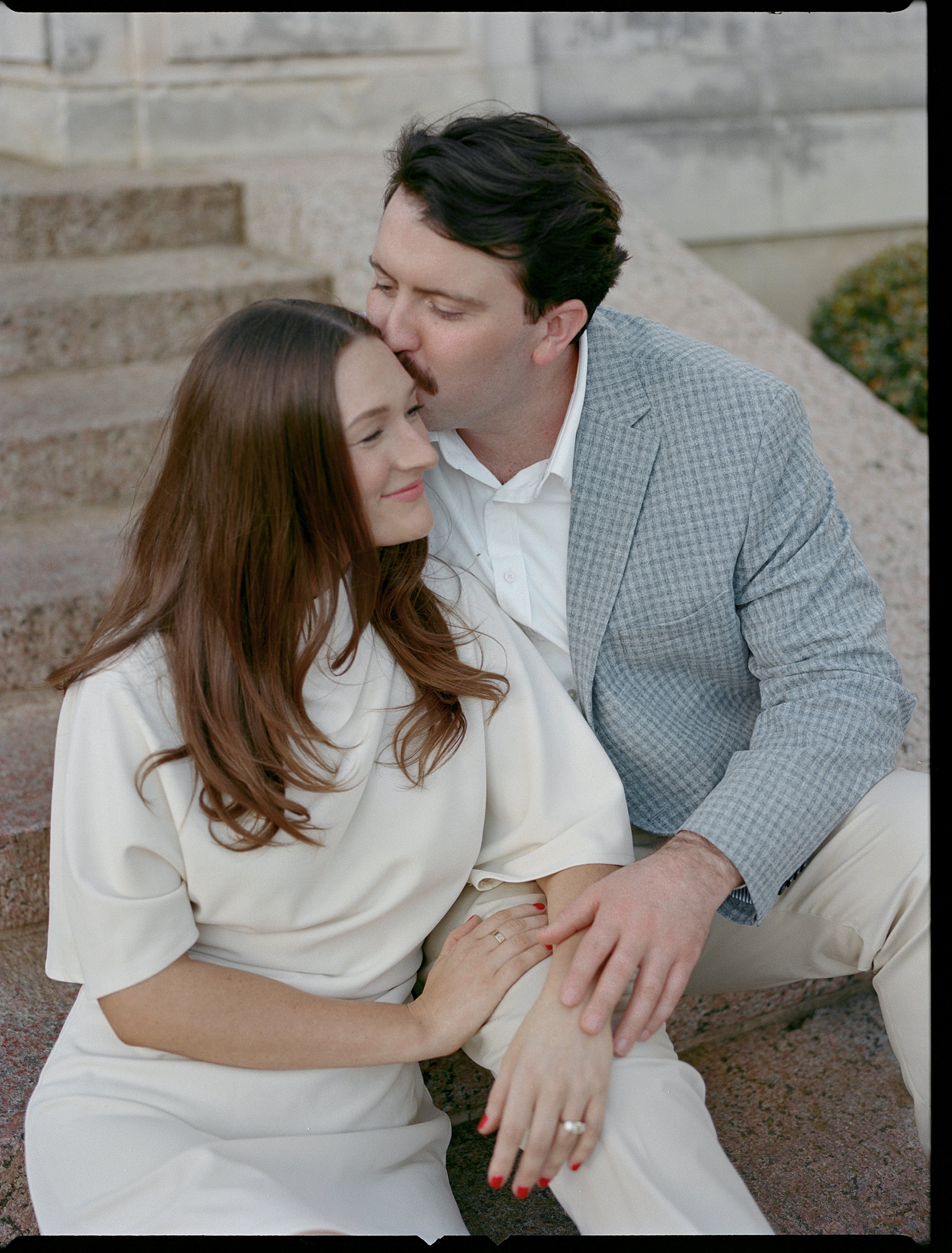 A man and woman sitting on outdoor stairs, with the man kissing the woman's forehead and holding her hand, both smiling.