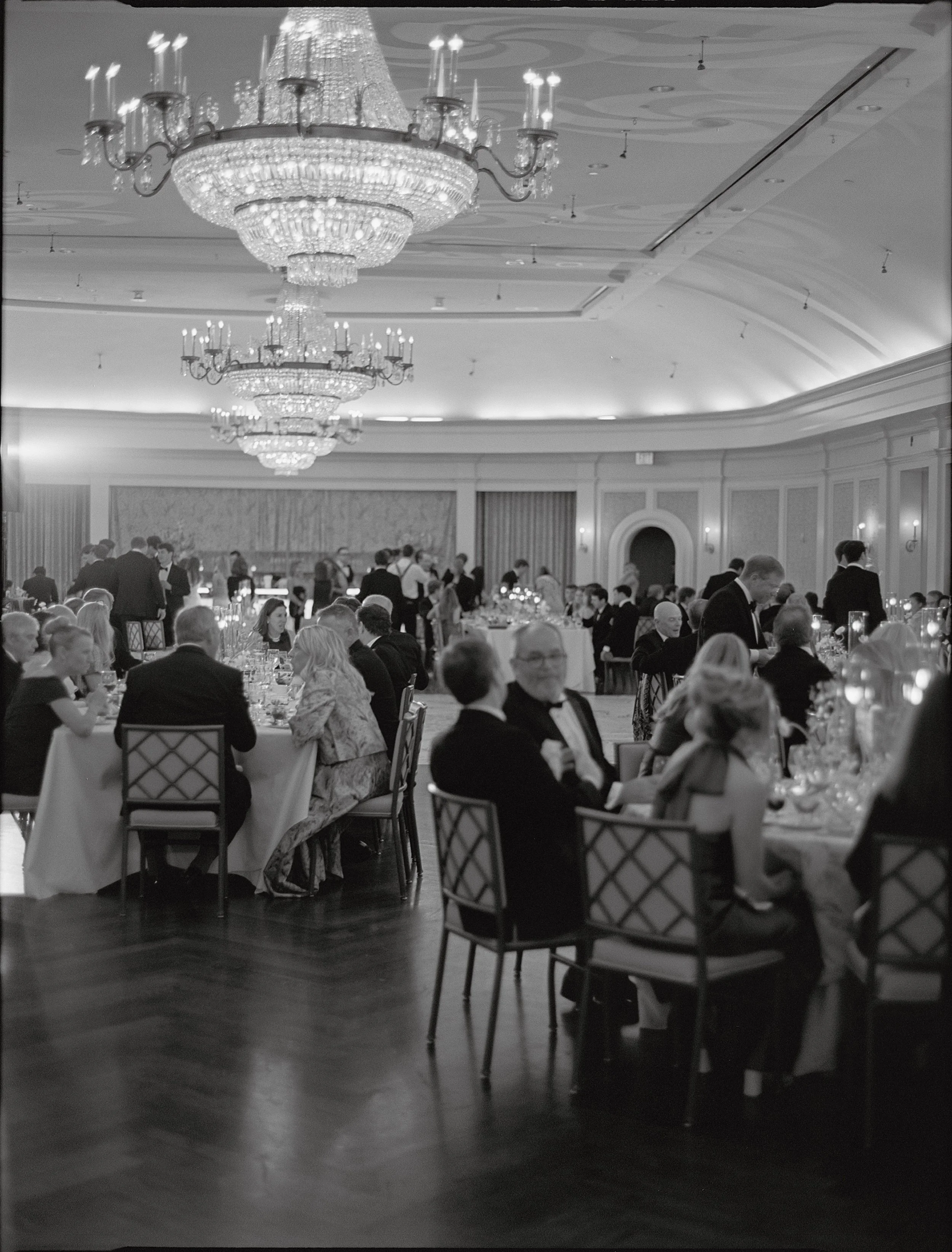 A black and white photo of an elegant banquet hall with chandeliers, filled with well-dressed guests seated at round tables, enjoying a formal event.