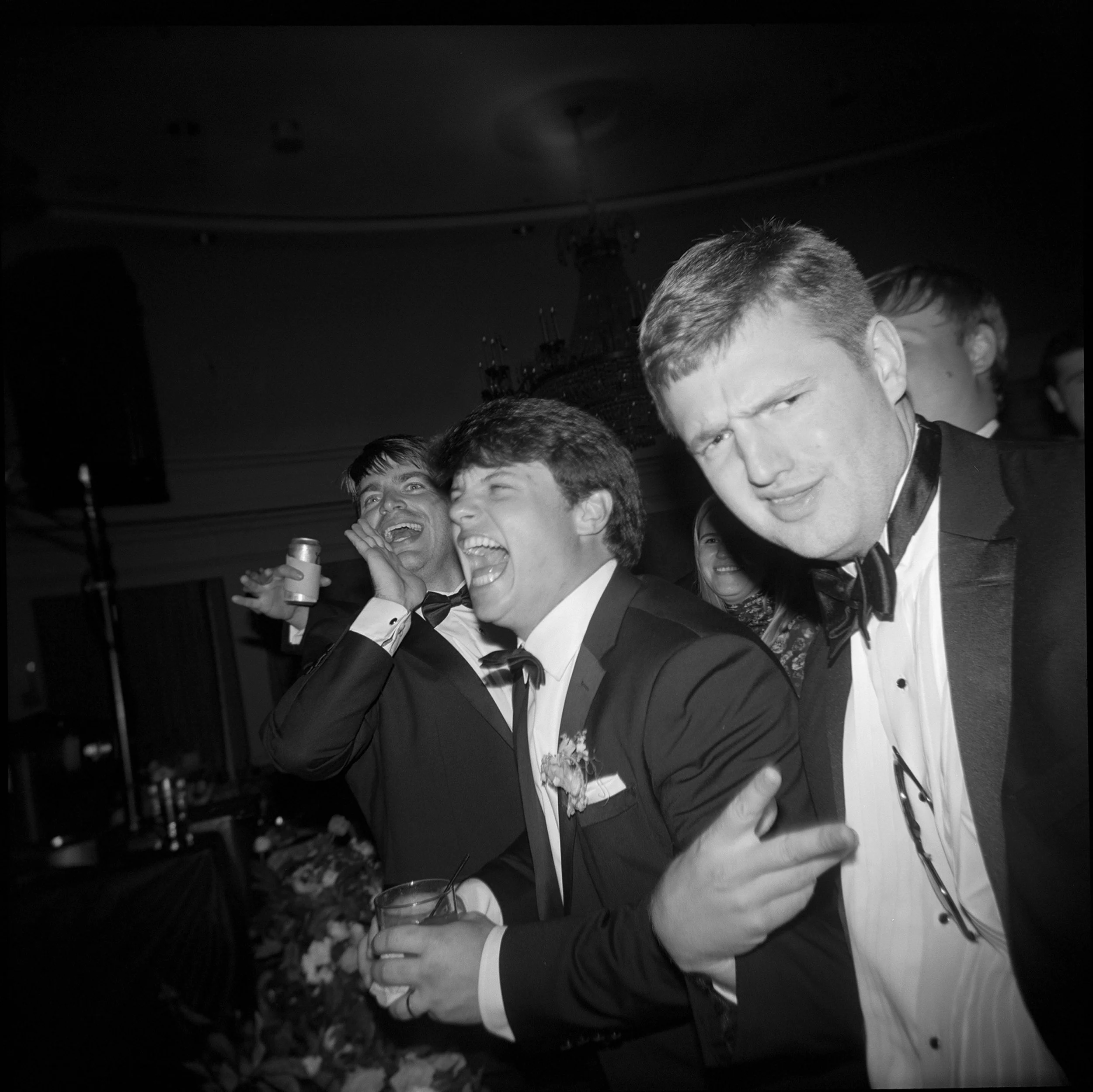 Black and white photo of young men in tuxedos celebrating at a formal event, laughing and posing with playful expressions.