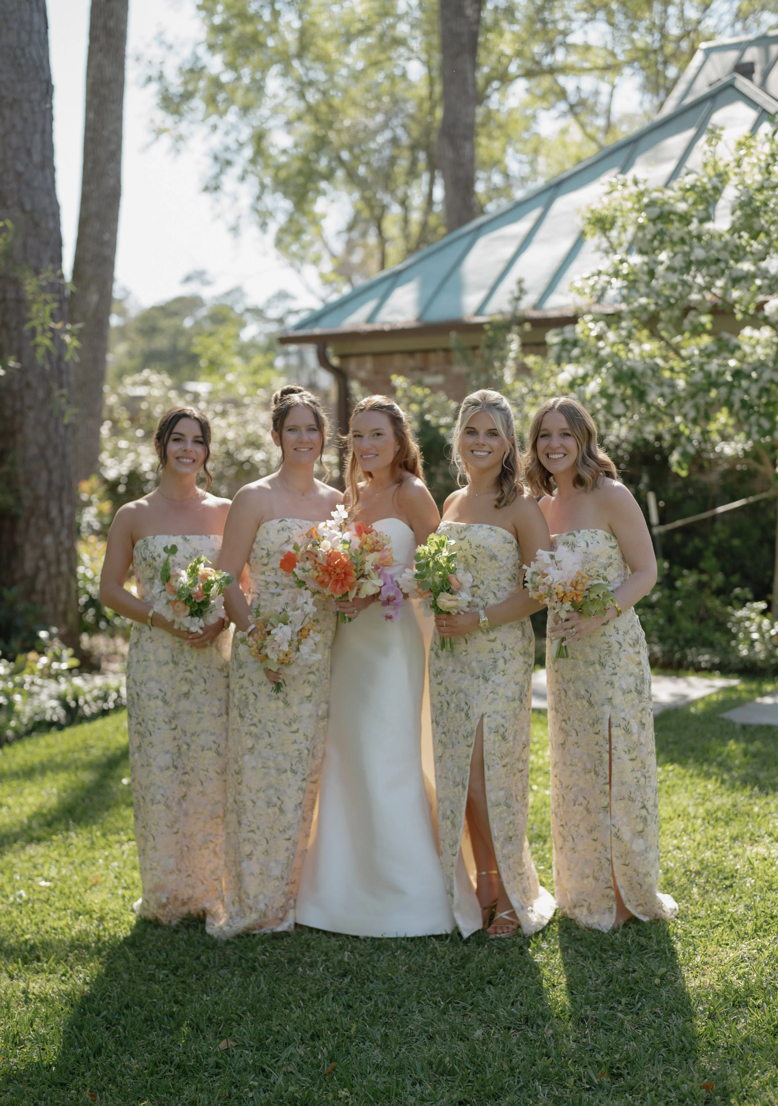 A bride and four bridesmaids standing outdoors on a grassy area during daytime. The bride is in a strapless white wedding gown holding a bouquet, with the bridesmaids in matching long floral dresses, each holding smaller bouquets. They are smiling, w