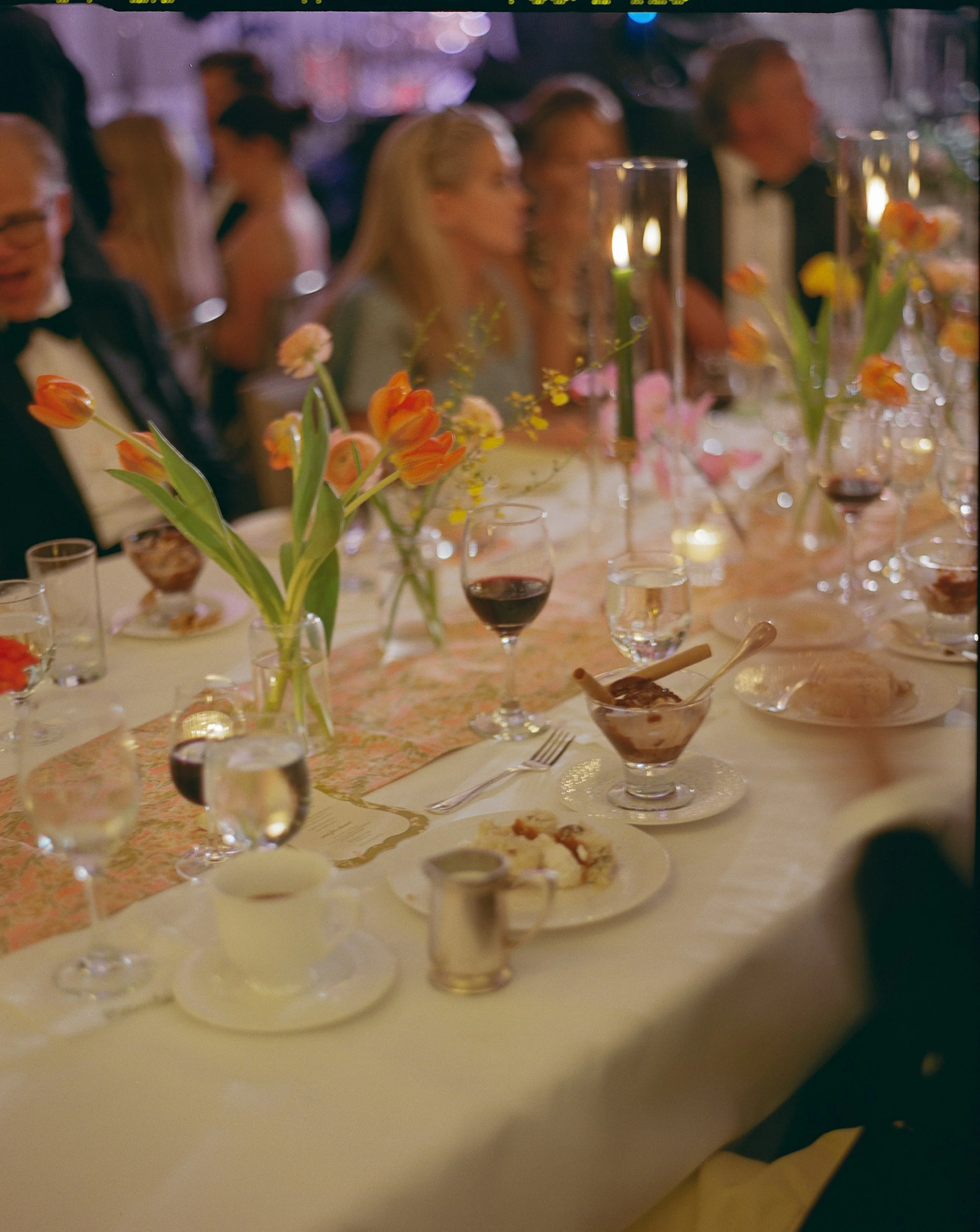 A formal dinner table set with pink and orange tulip centerpieces, glasses of red wine and water, plates with desserts, and lit candles, with people dressed in elegant attire sitting and talking.
