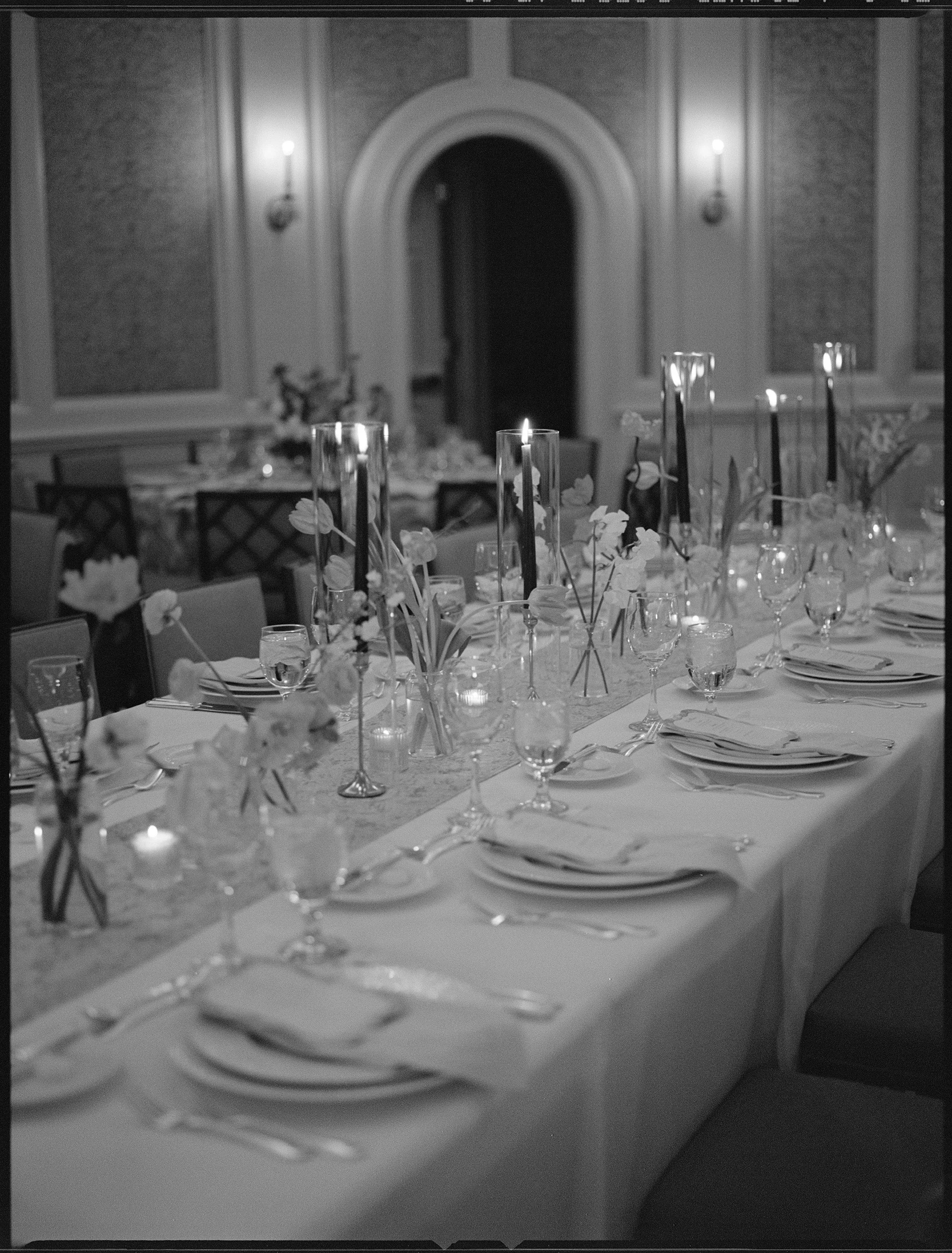 Elegant banquet table set with plates, glasses, silverware, floral centerpieces, candles in glass holders, and tall candles in vases, in a decorated event hall.