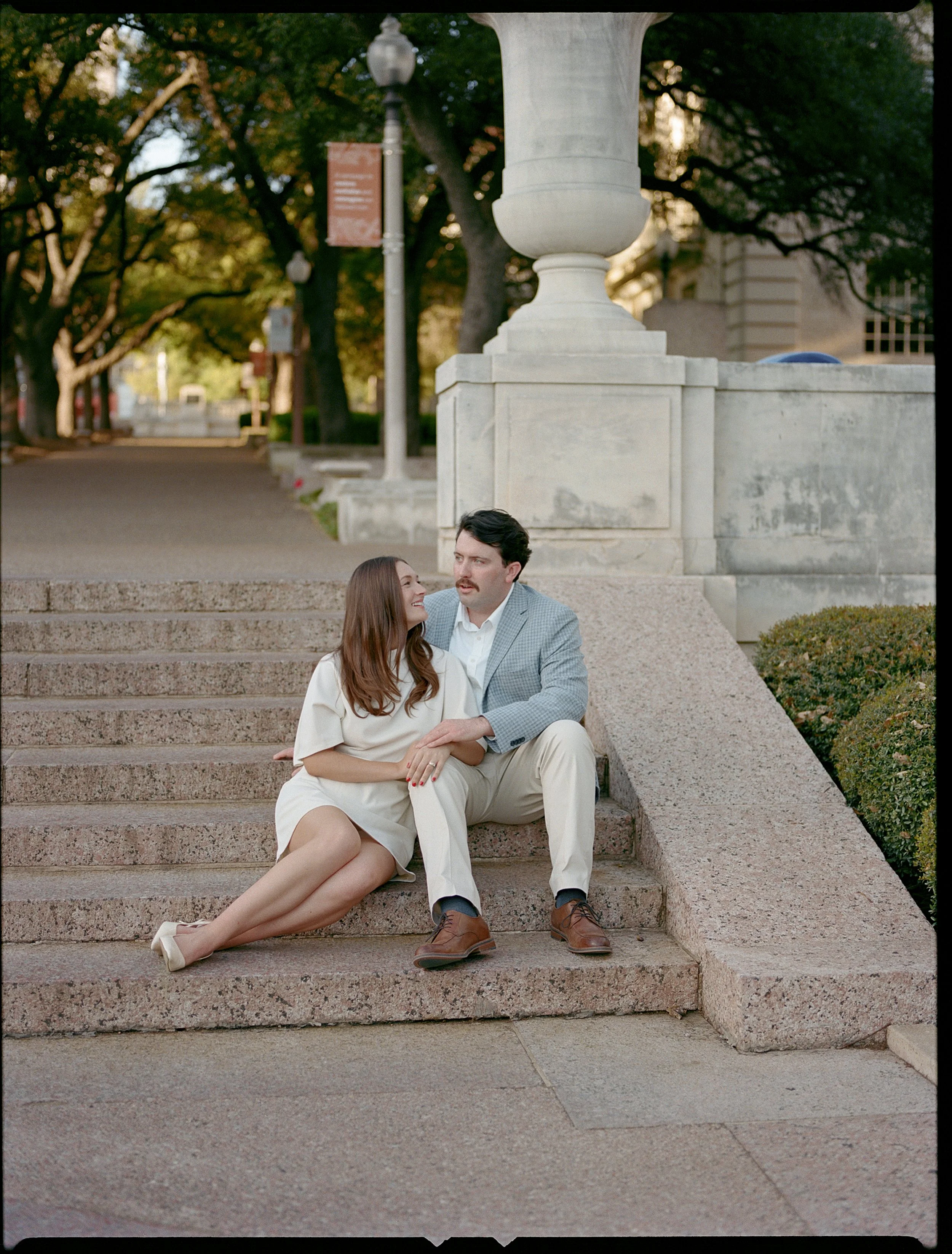 A couple sitting on stone steps outdoors, near a large stone urn and surrounded by trees and greenery.