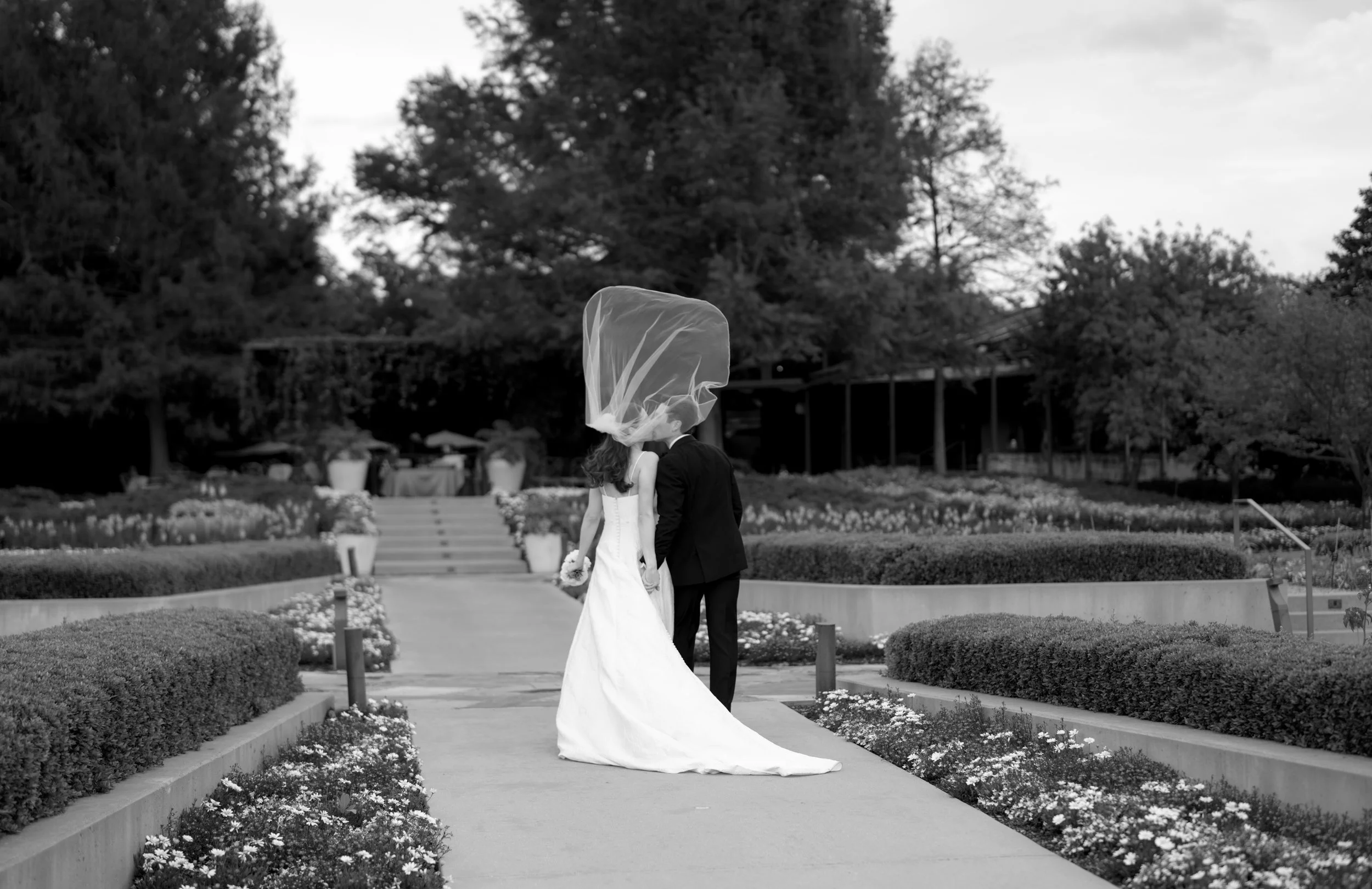 A black and white photograph of a bride and groom kissing on a garden path, with the bride in a white wedding dress holding flowers, and the groom in a dark suit, with a veil blowing in the wind.