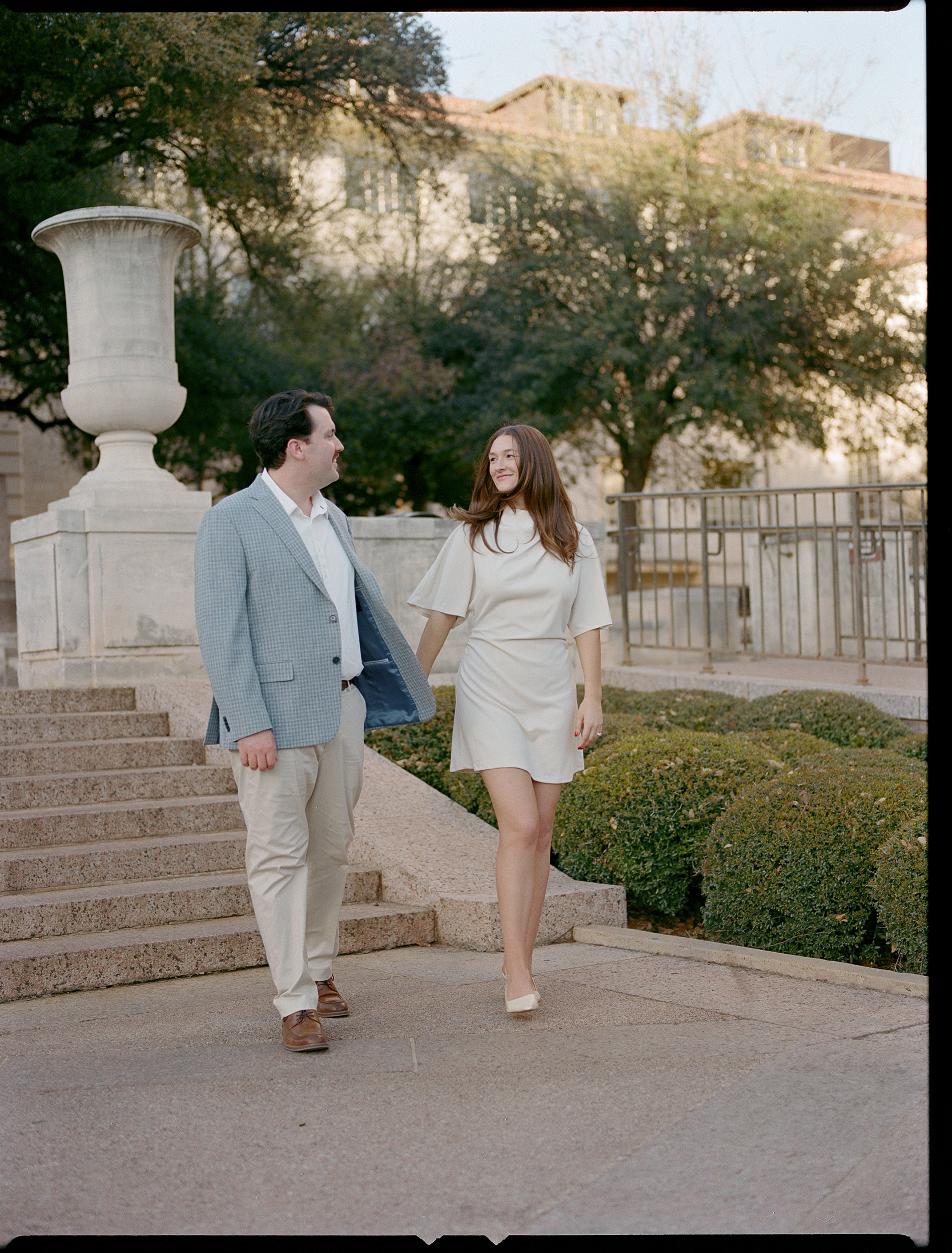 A man and woman holding hands and walking up the stairs outdoors, near a large decorative urn and trees with a building in the background.