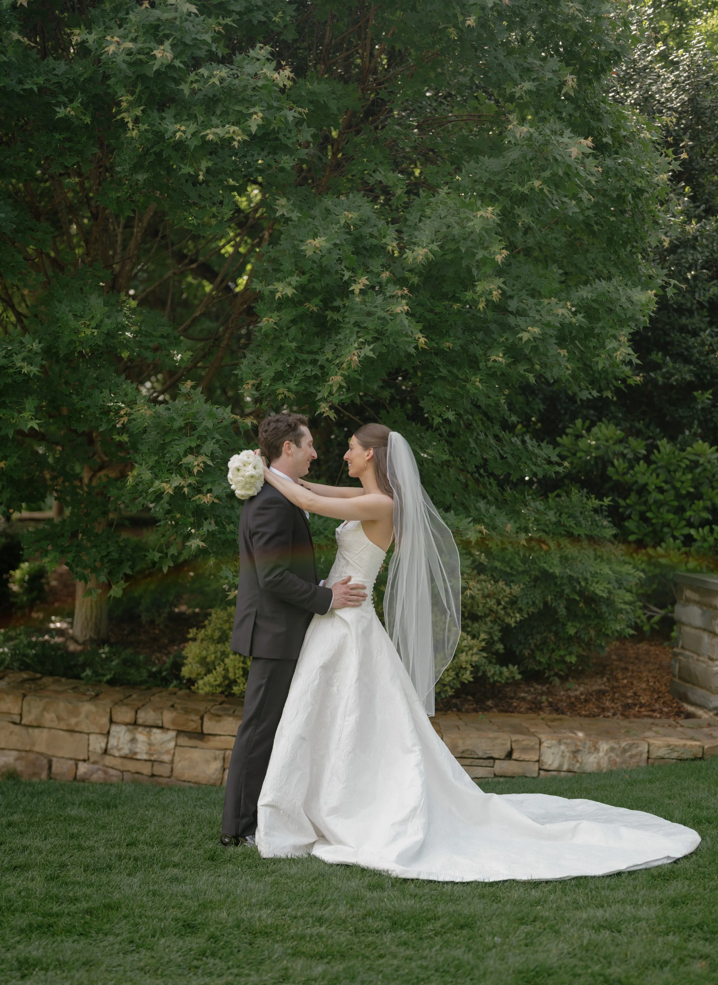 A bride and groom standing outdoors on green grass, embracing in front of a large leafy tree during their wedding.