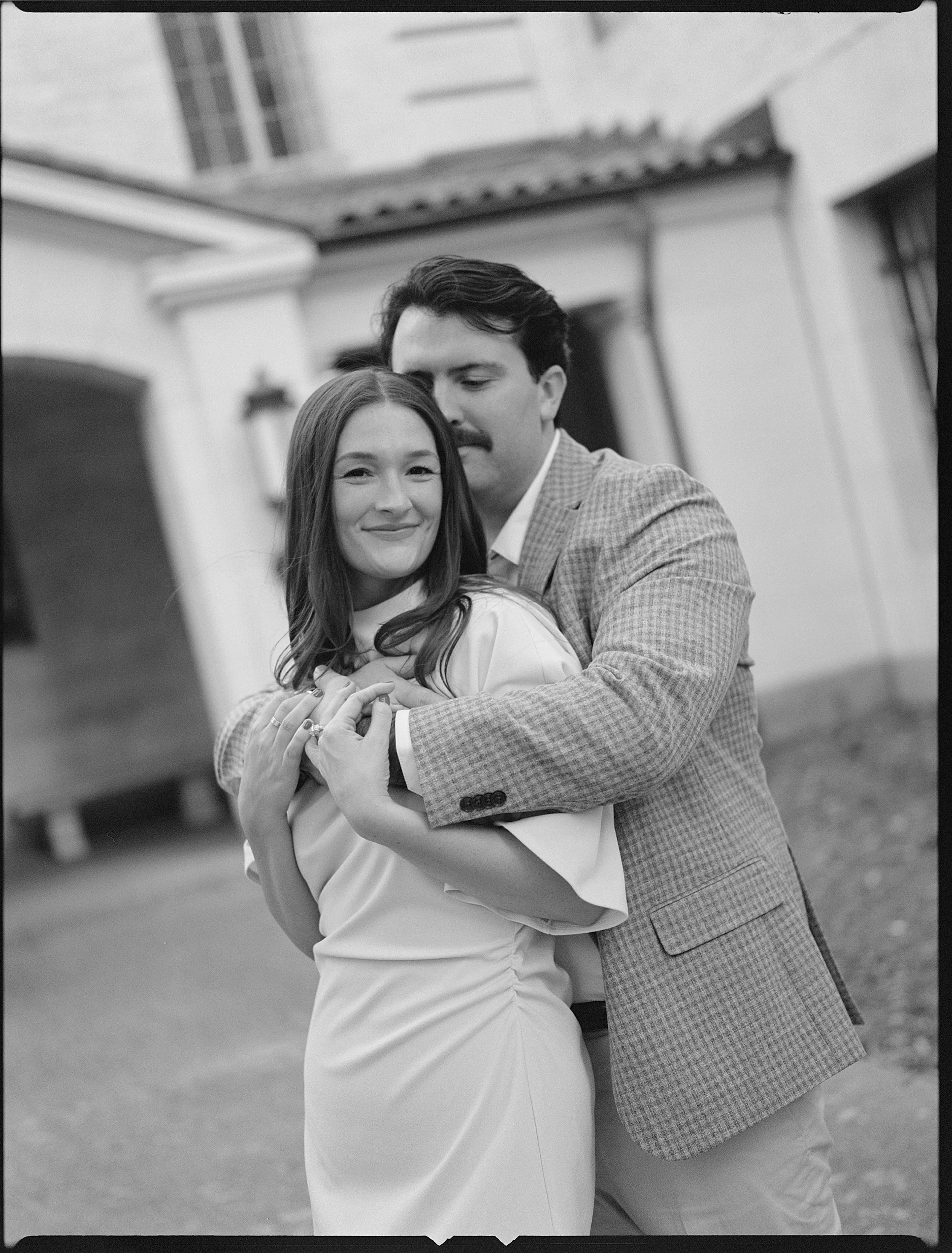 A black and white photo of a happy couple embracing outside in front of a building, with the man holding the woman from behind, both smiling.