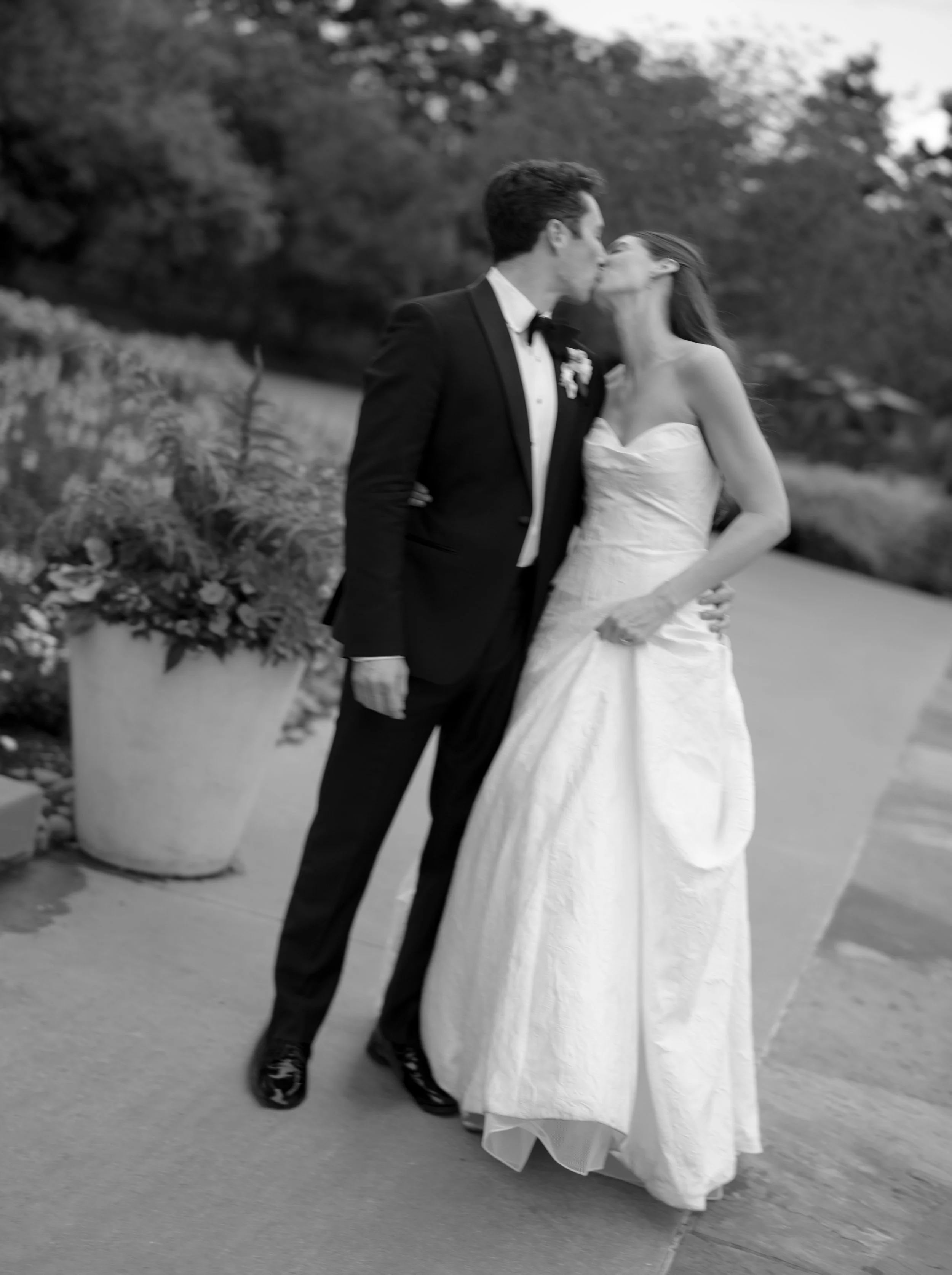 A black and white photo of a bride and groom kissing outdoors, with trees and plants in the background, on a paved walkway.