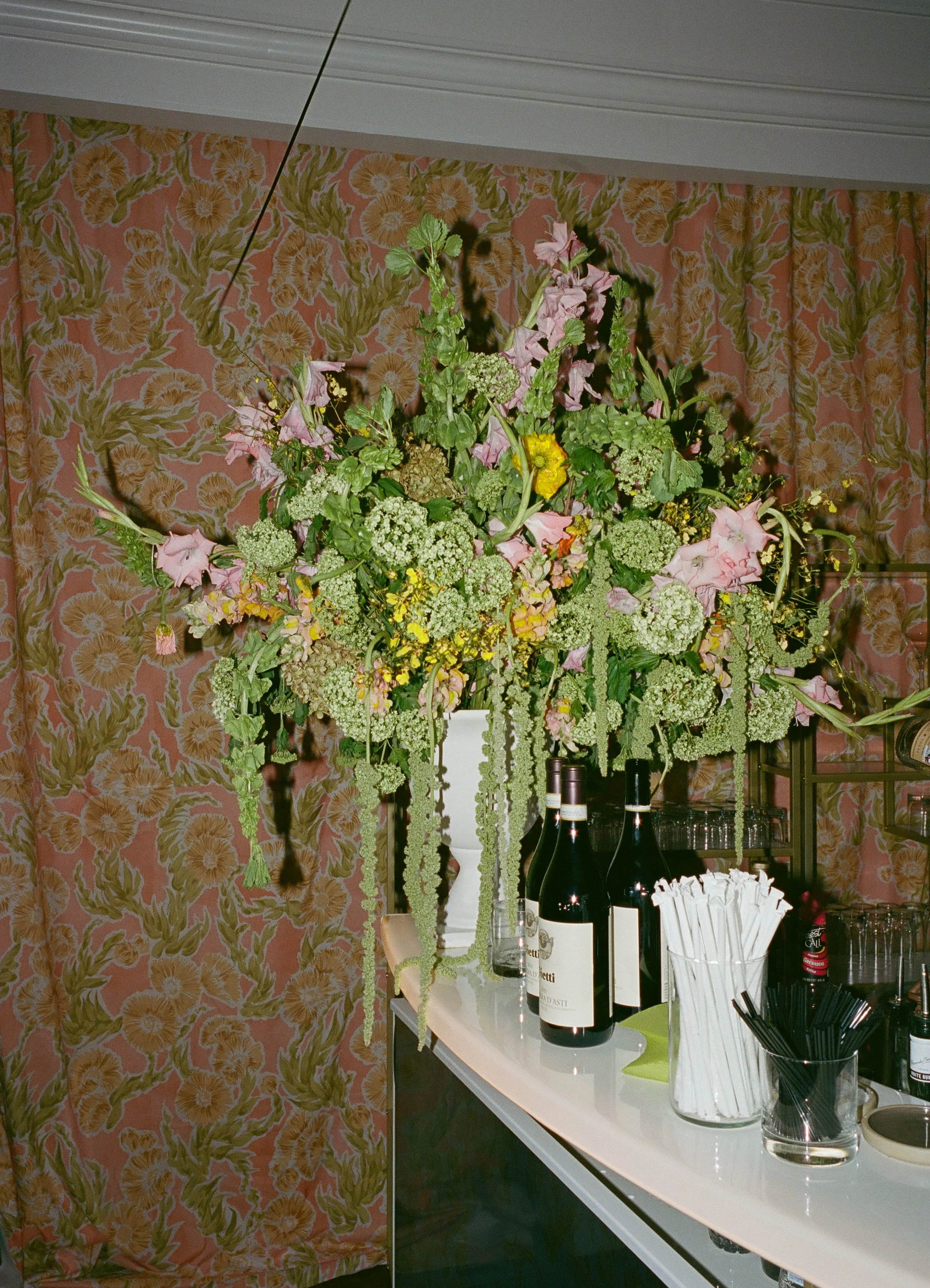 A large floral arrangement in a white vase on a bar counter, with wine bottles, straws, utensils, and glasses nearby, against a pink and yellow patterned floral wall.