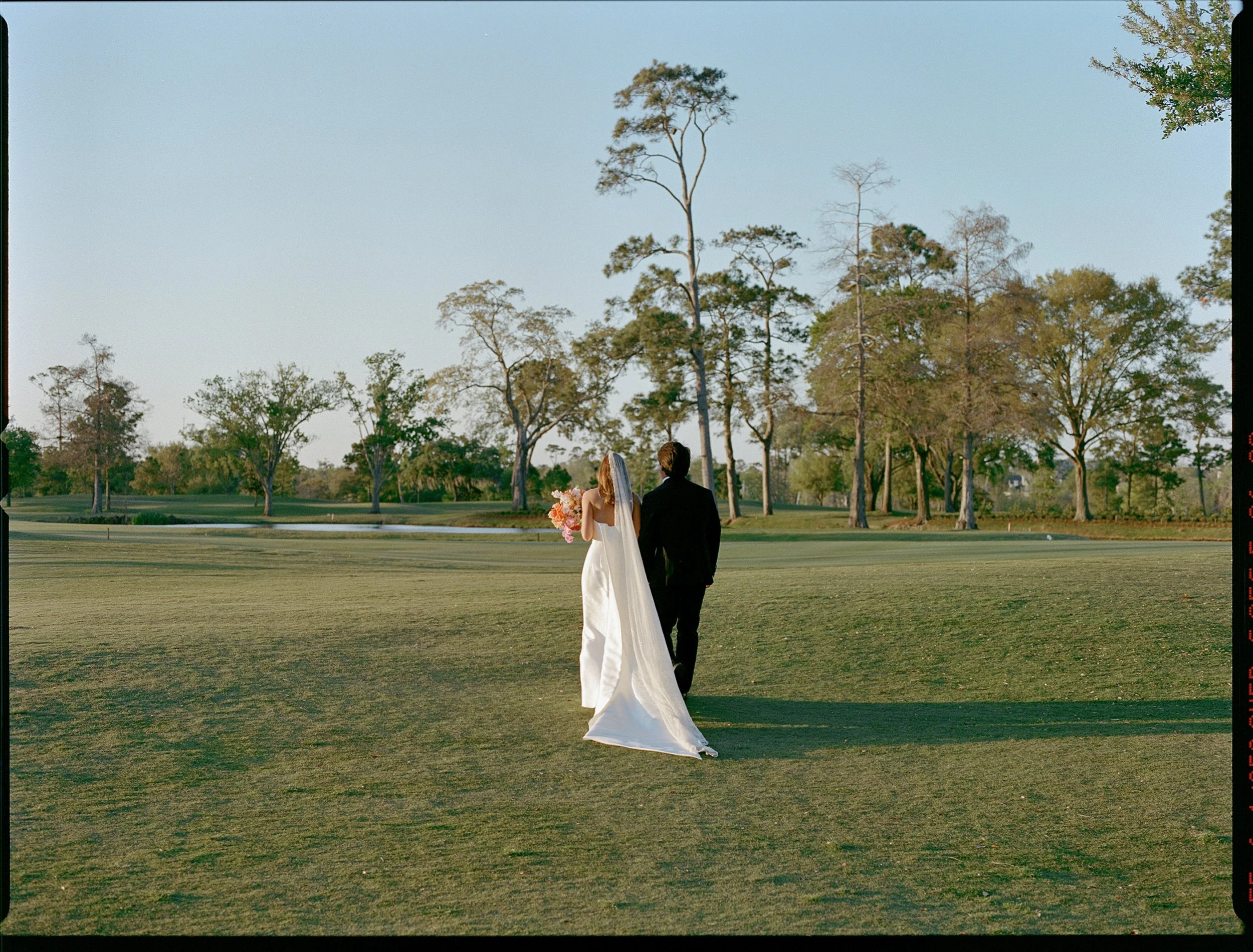 A bride with a bouquet and a groom in a suit walking together on a golf course with trees and a pond in the background.