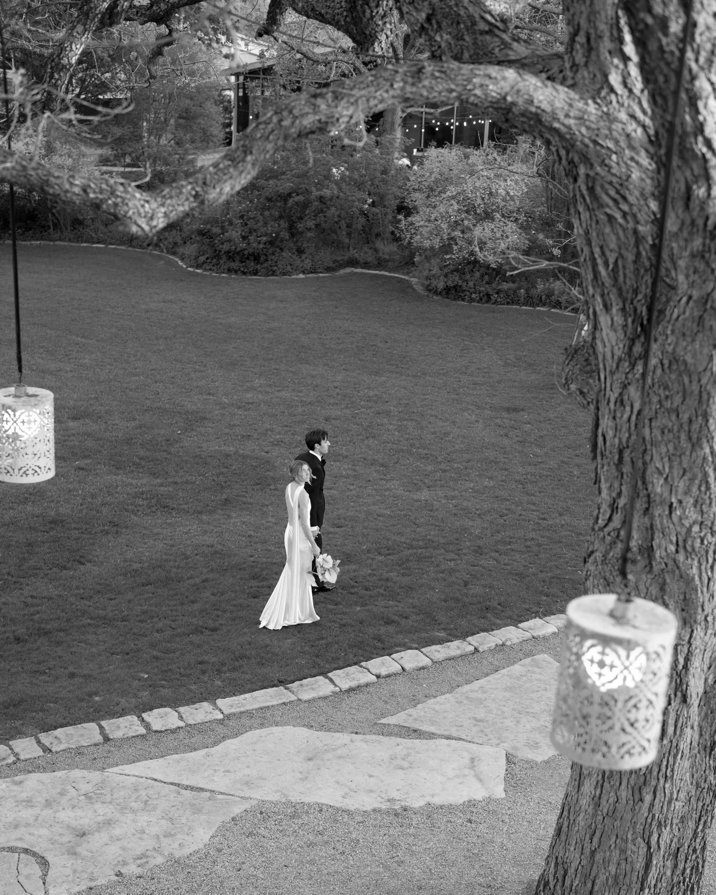 A bride and groom standing on a lawn at an outdoor wedding, captured through a tree with hanging lanterns.