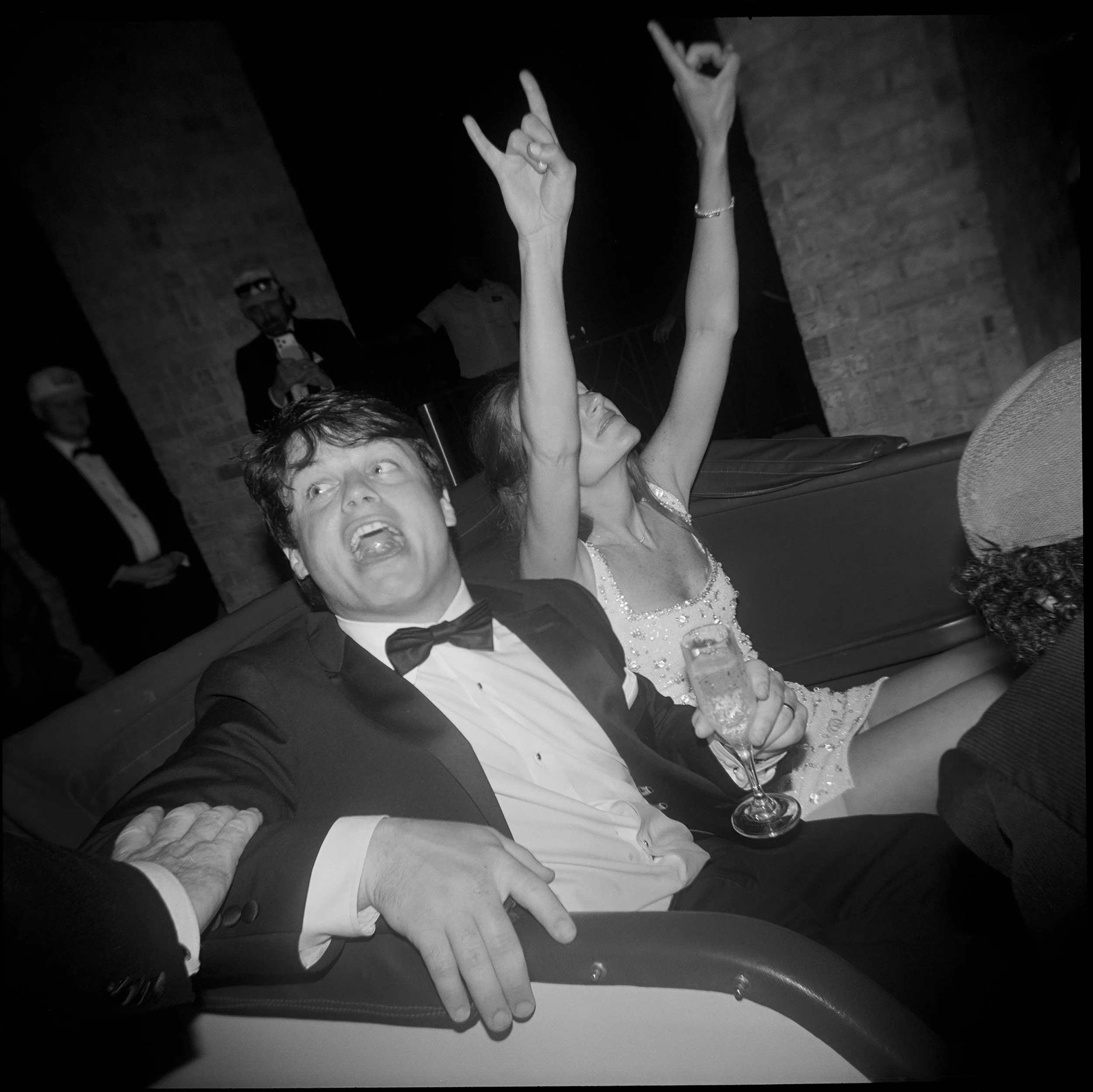 A black and white photo of a man in a tuxedo with a bow tie and a woman in a dress, both sitting on a sofa, celebrating with drinks. The woman is raising her arms, and the man is looking up. There are people in the background, some wearing tuxedos, a