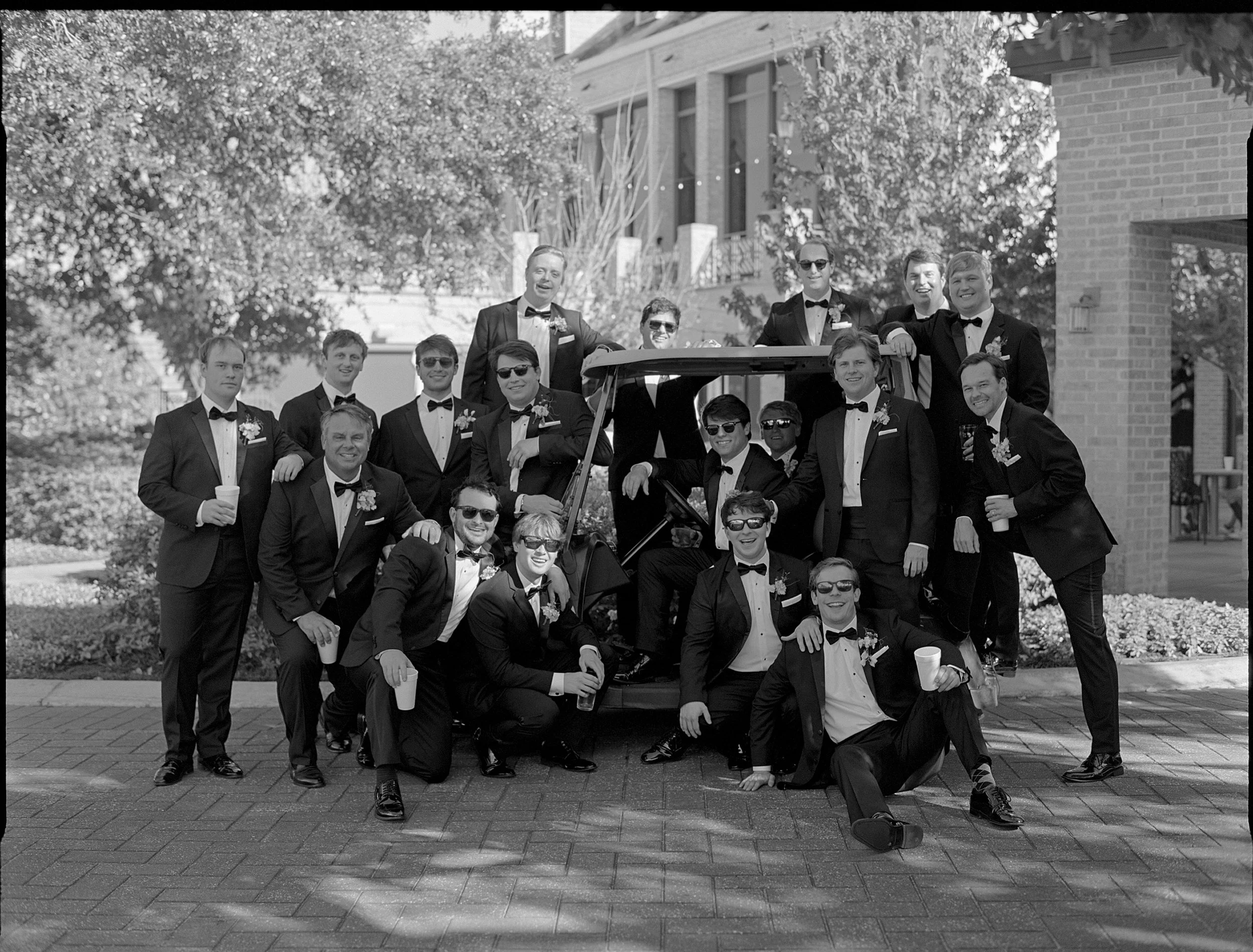 A group of men dressed in tuxedos and sunglasses posing around and on a golf cart outdoors during daytime.