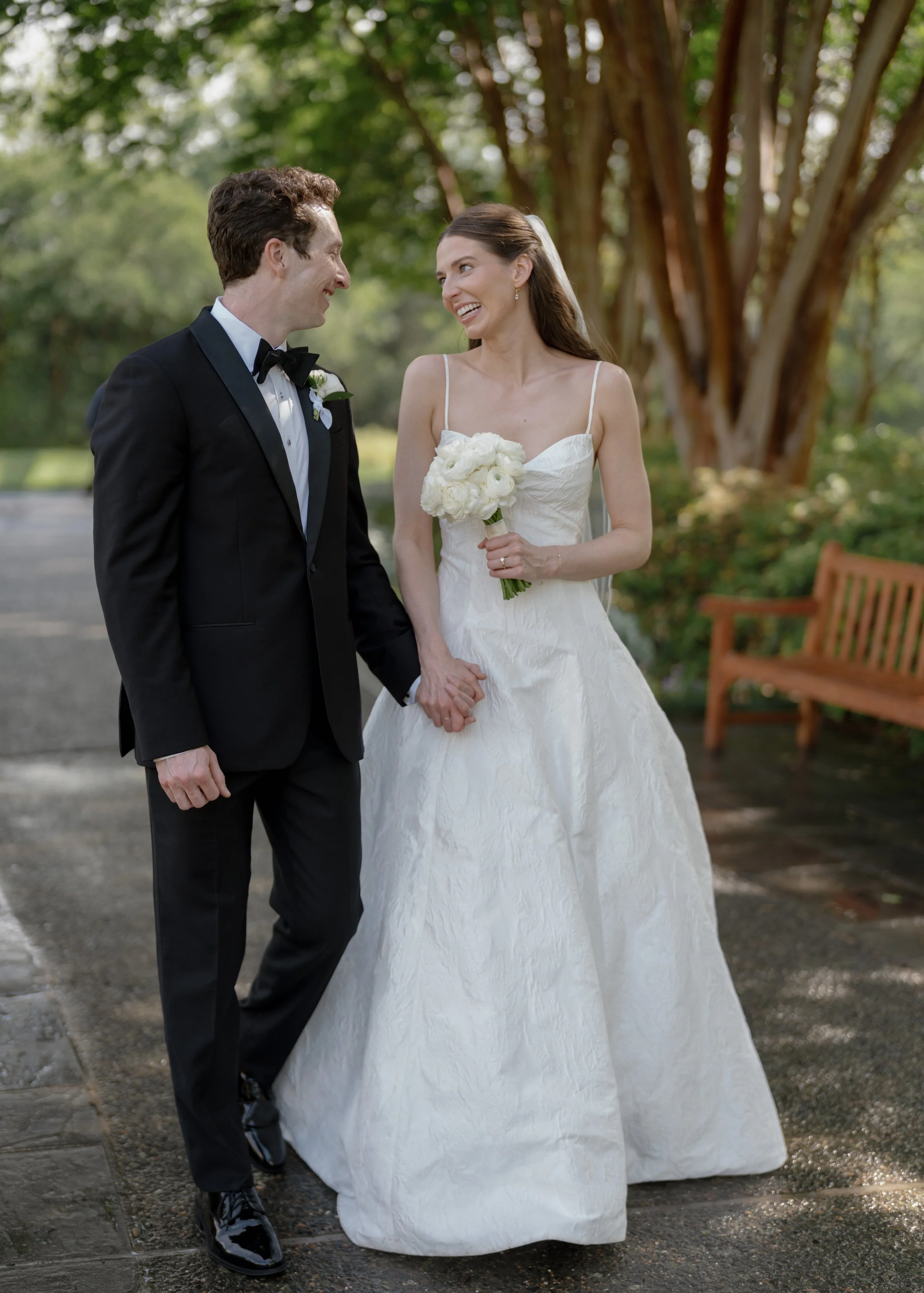 A bride and groom holding hands and smiling at each other at the Dallas Arboretum, with trees and a bench in the background.
