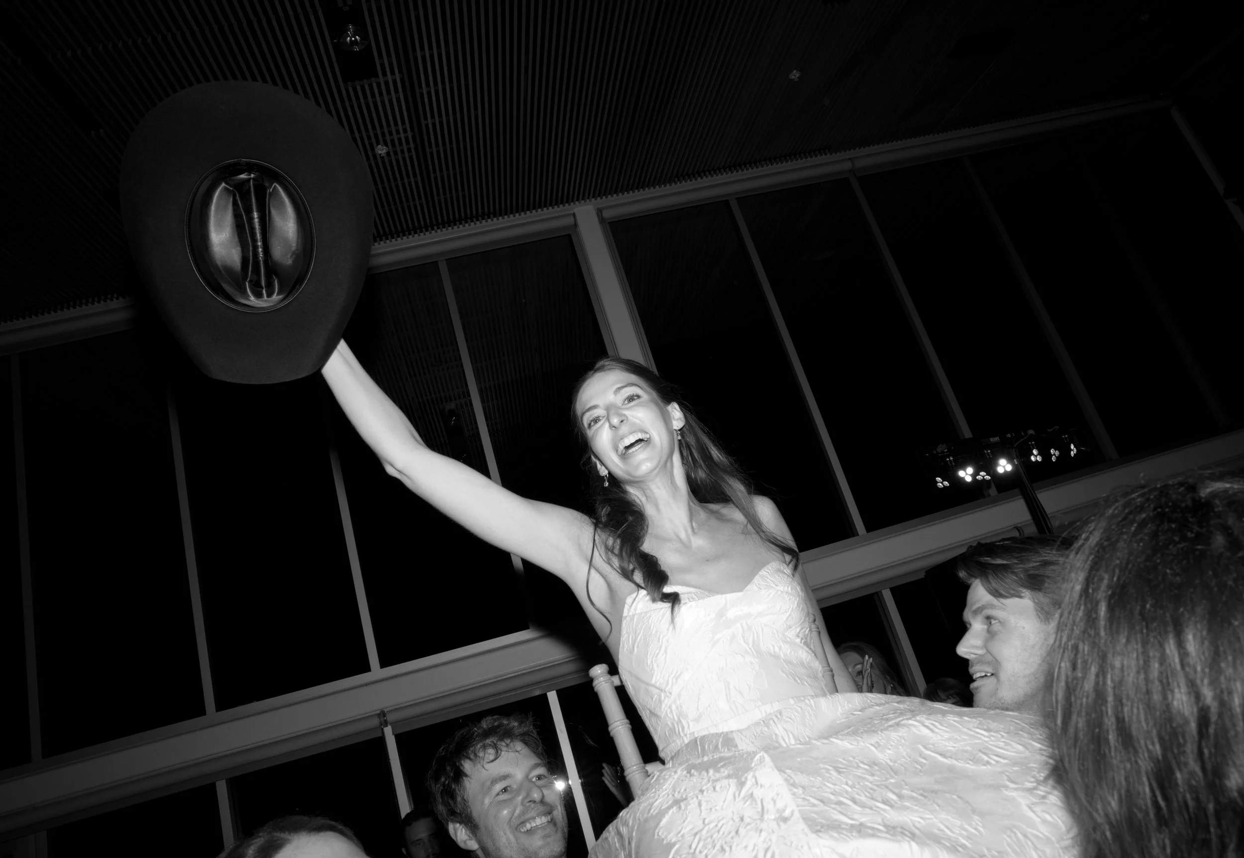 Bride in wedding dress holding a hat high in the air during celebration, surrounded by smiling guests in an indoor venue.