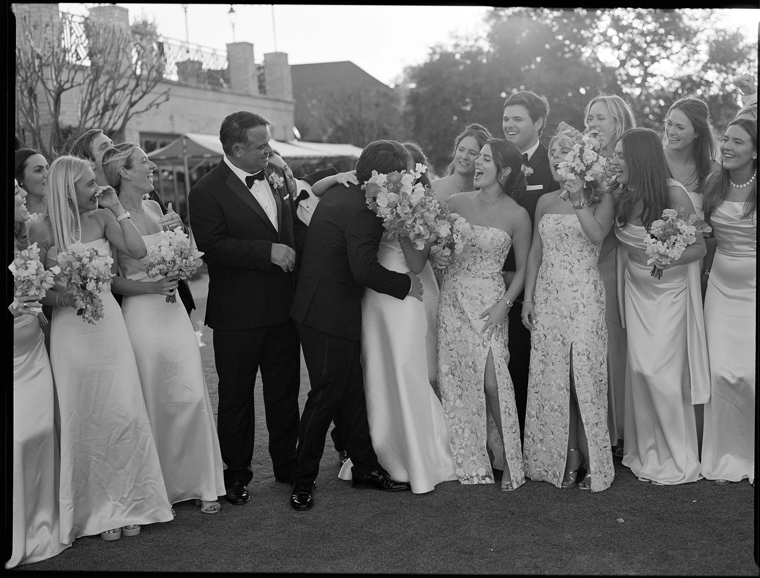 A group of people at a wedding, with the bride and groom kissing in front of the wedding party, all smiling and holding flowers.