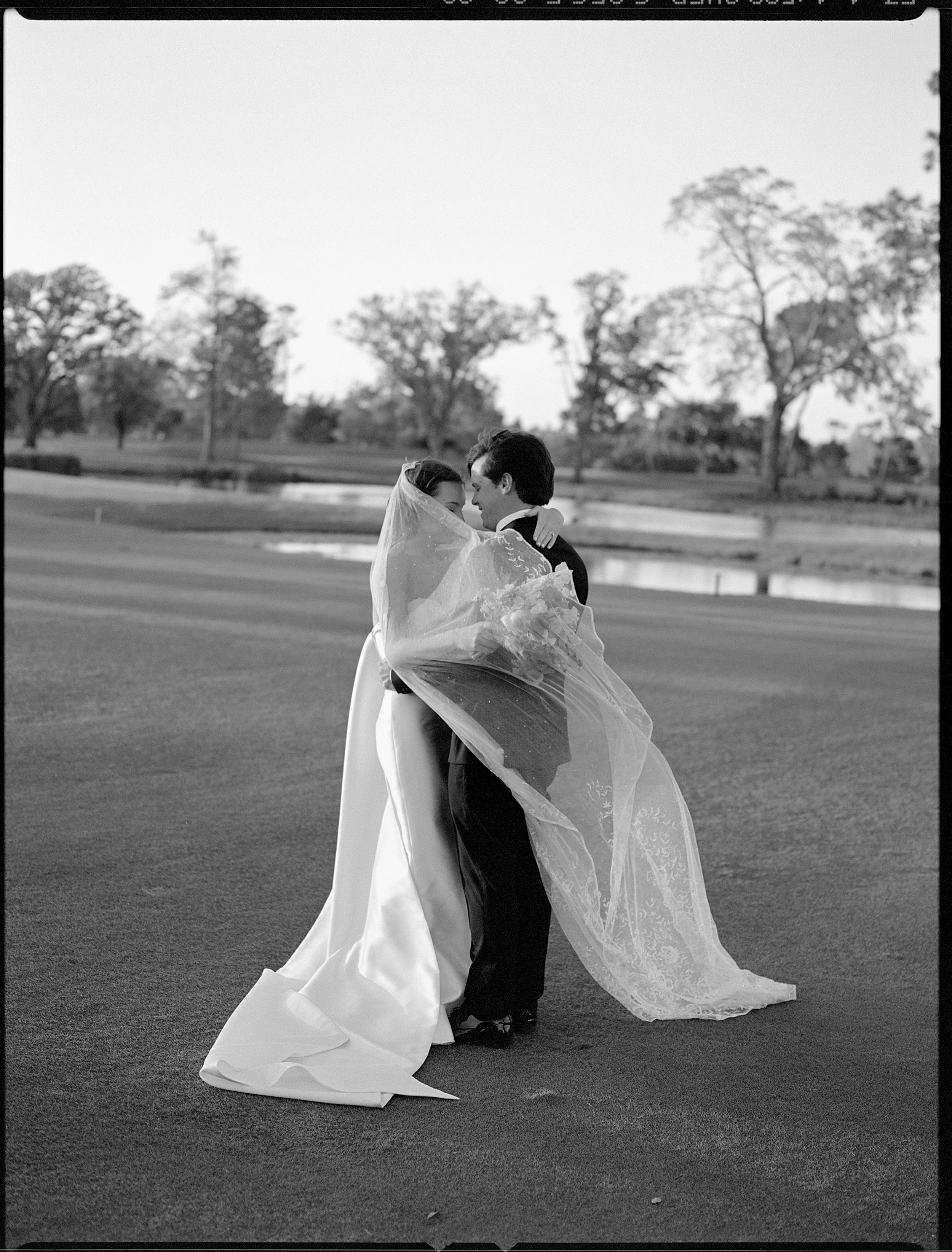 A bride and groom embrace on a paved outdoor area in black and white, with water and trees in the background.