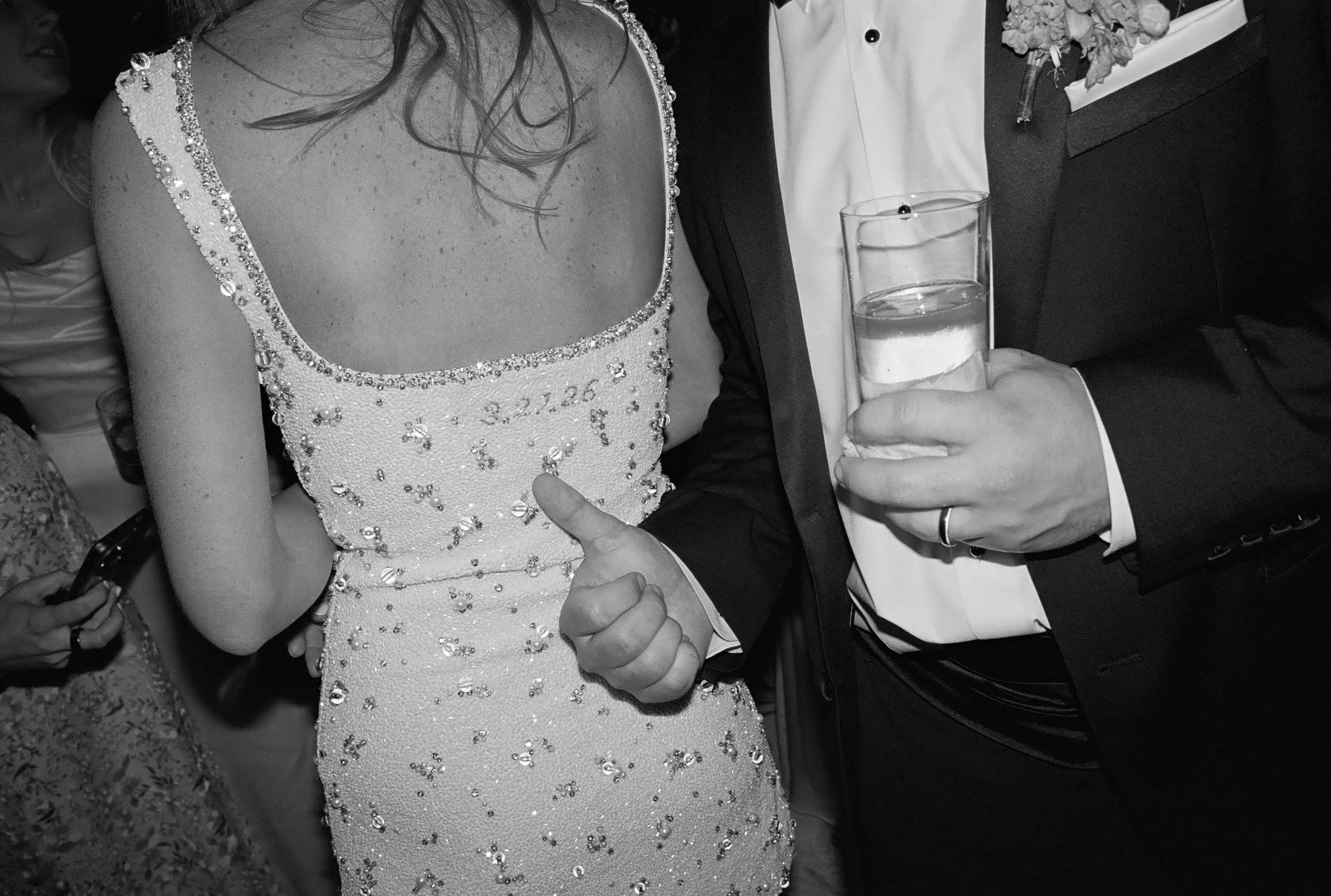 A person in a tuxedo holding a glass of water with a ring on their finger, standing next to a woman in a sequined dress with the date 3.22.26 embroidered on it, at a formal event.