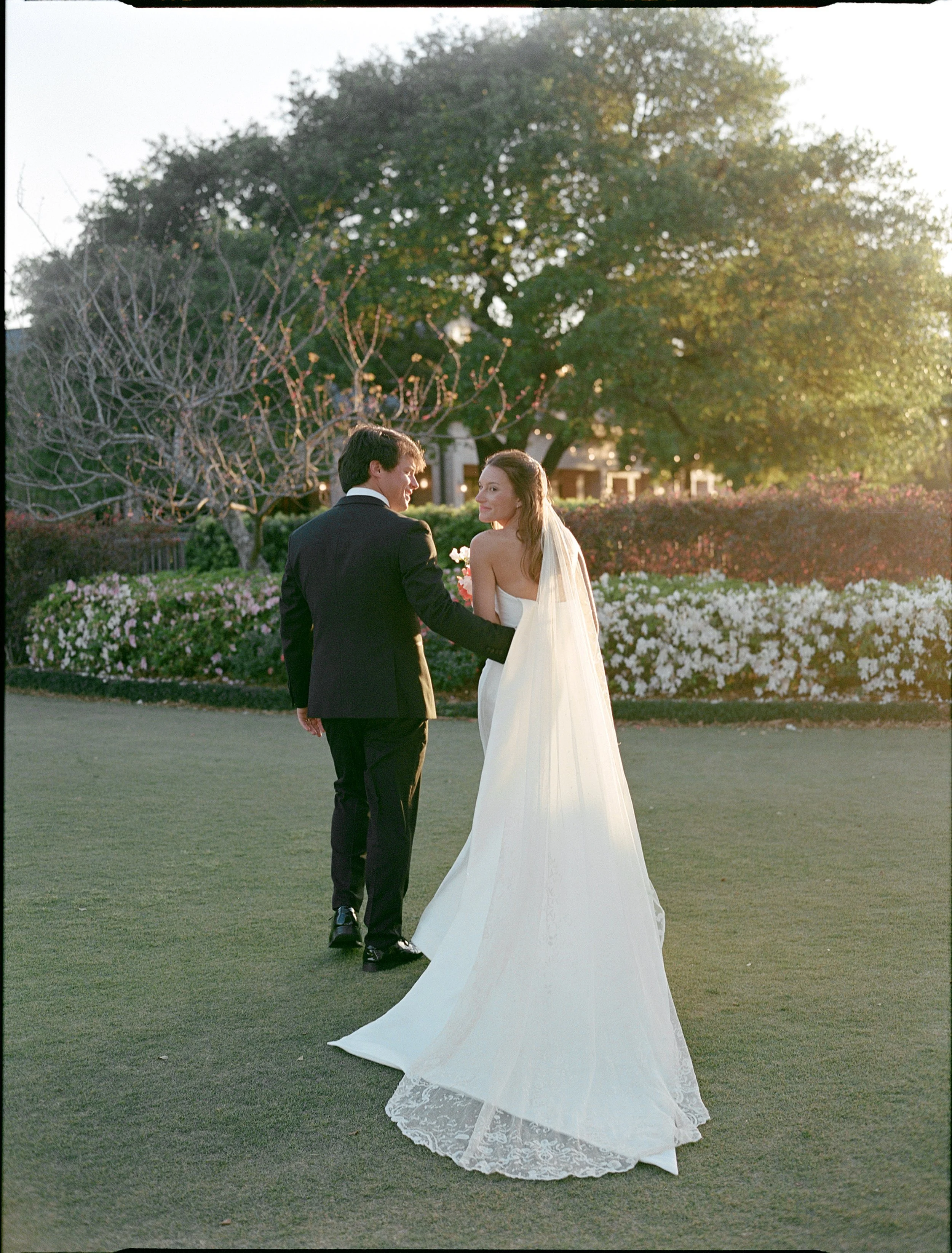 A bride and groom walking together outdoors during sunset, with lush green trees and flowers in the background.