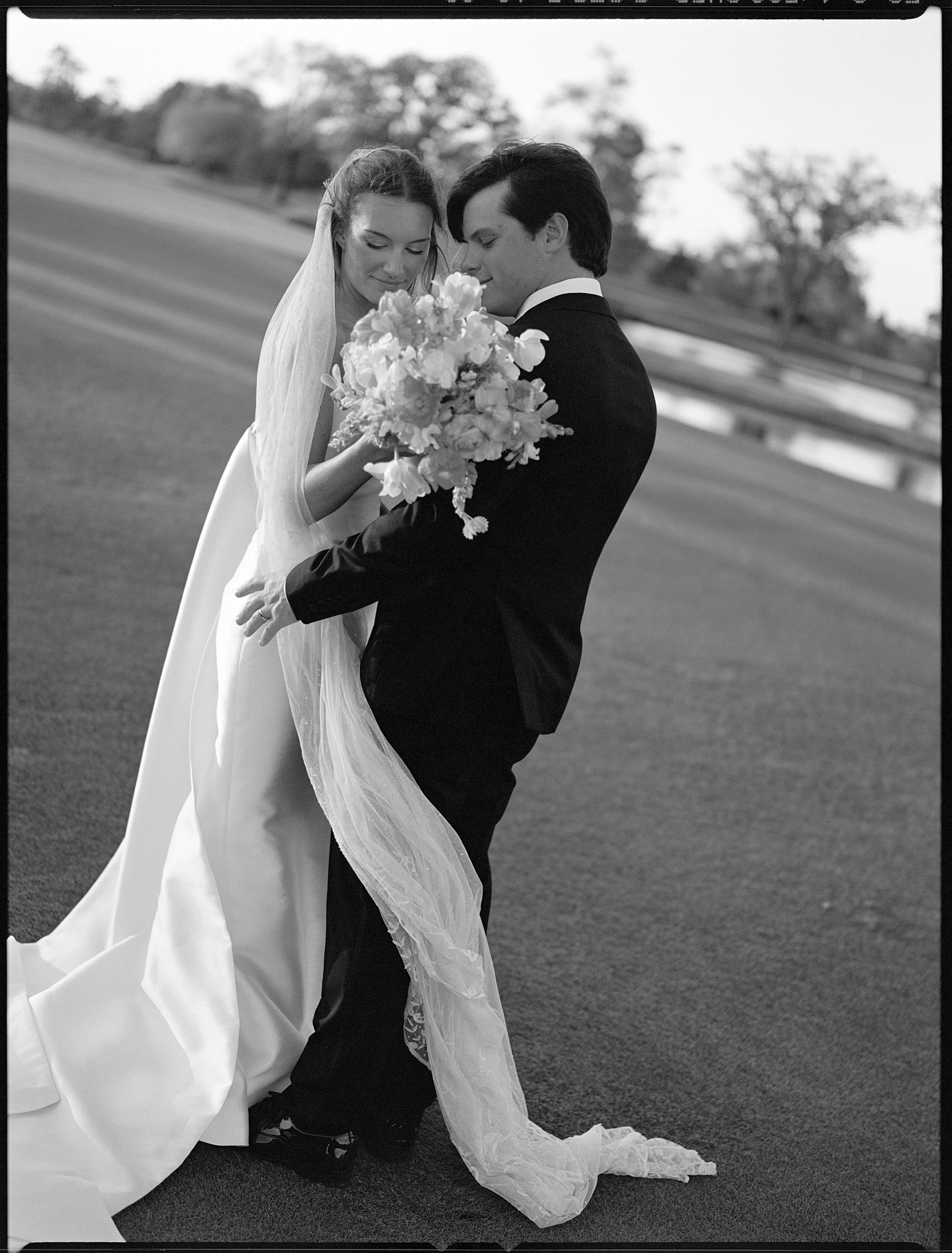 Black and white photo of a bride and groom on a grassy field, with the groom holding a bouquet of flowers as they share an intimate moment at River Oaks Country Club