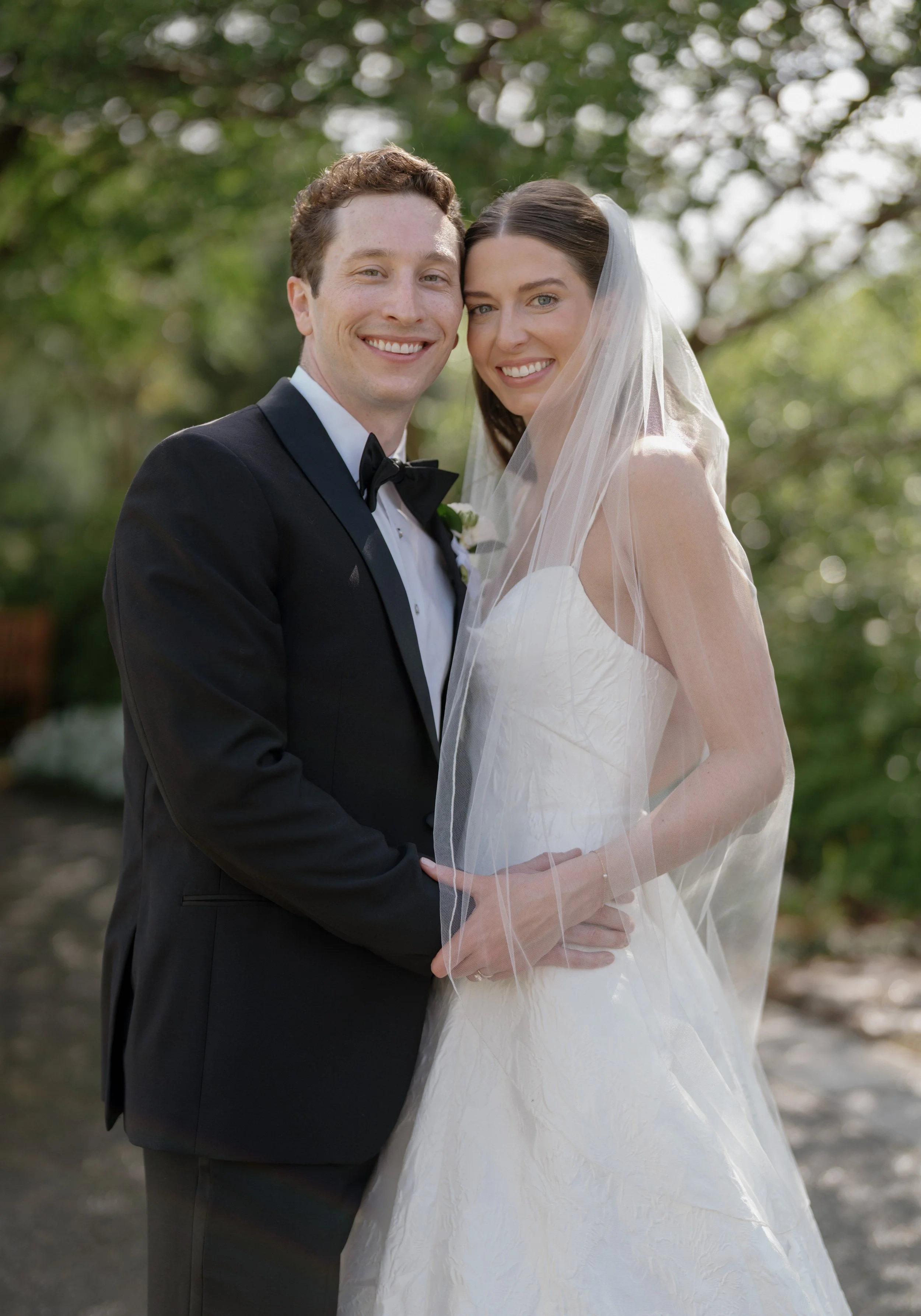 A bride and groom in wedding attire smiling at the Dallas Arboretum, with trees and greenery in the background.