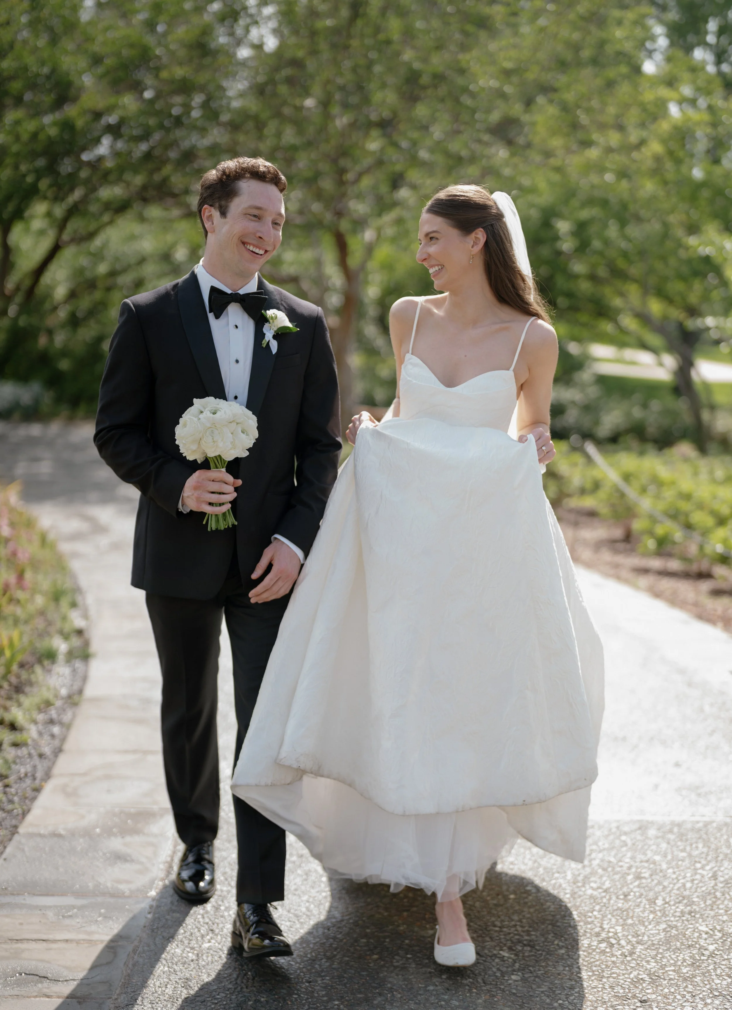 A smiling bride in a white wedding gown and a smiling groom in a black tuxedo, holding a bouquet of white flowers, walking outdoors on a sunny day with trees in the background.