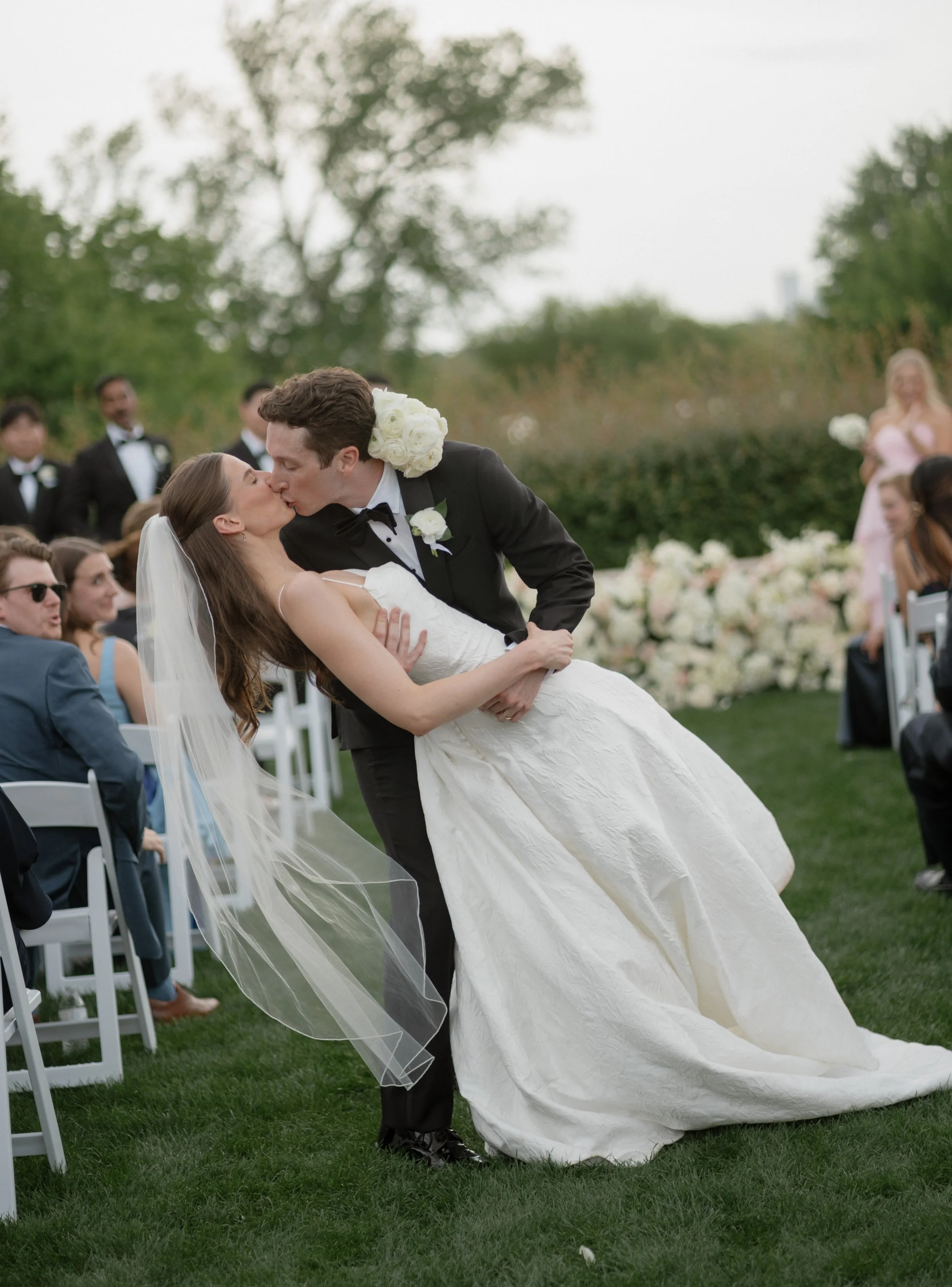 A bride and groom kissing during their outdoor wedding ceremony, with bridesmaids, groomsmen, and guests seated around them.