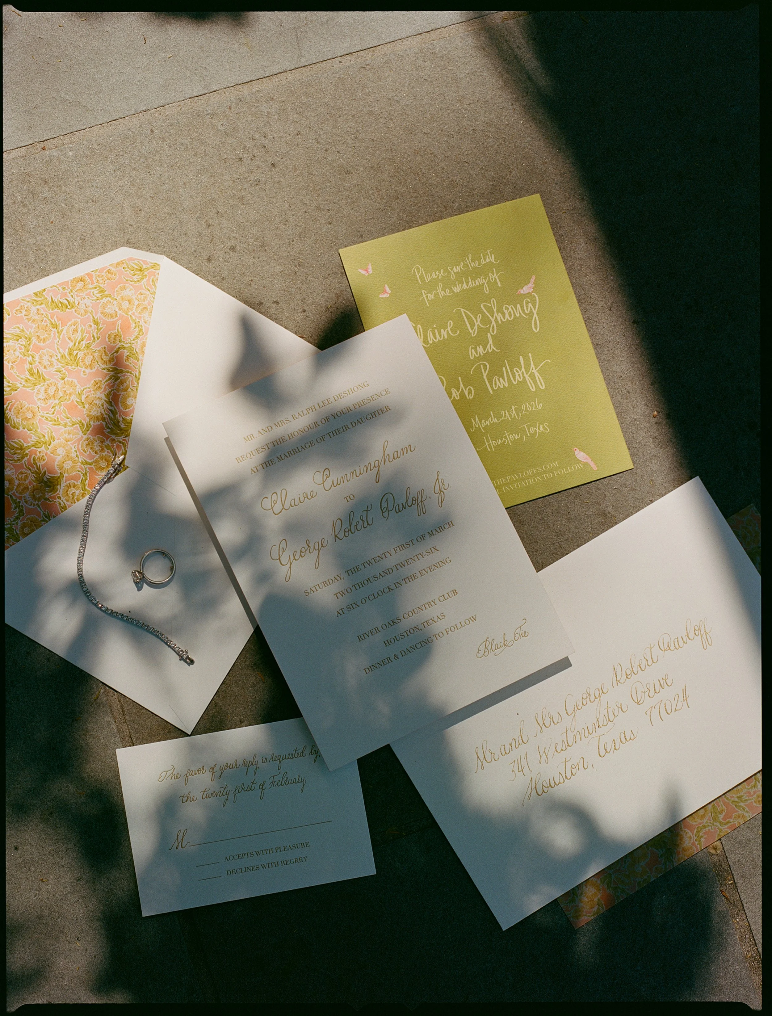 Wedding invitation suite including an envelope, invitation card, RSVP card, and additional card, with a ring and decorative paper, all arranged on an outdoor surface with shadows.