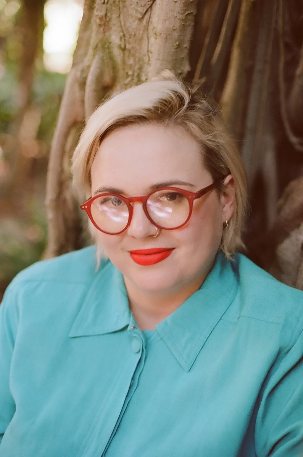 A woman with short blonde hair and red glasses, wearing a teal blouse, standing outdoors near a tree with a background of blurred greenery.
