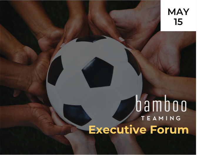 Two relay runners in yellow and blue athletic gear pass a baton on an outdoor track, mid-handoff, with one runner smiling. Mountains and green fields are visible in the background under an overcast sky.