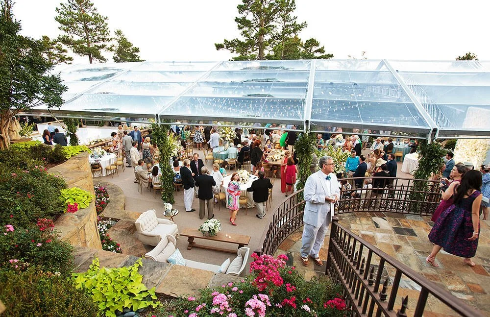 Outdoor event with guests socializing under a large transparent tent, surrounded by lush greenery and flowers.