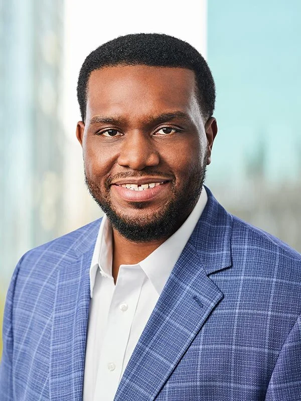 Professional headshot of a smiling man with short hair and a neatly trimmed beard, wearing a blue plaid blazer and white shirt, set against a blurred background