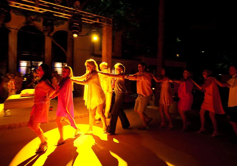 Group of people dancing together outside, illuminated by warm lighting in a lively atmosphere.