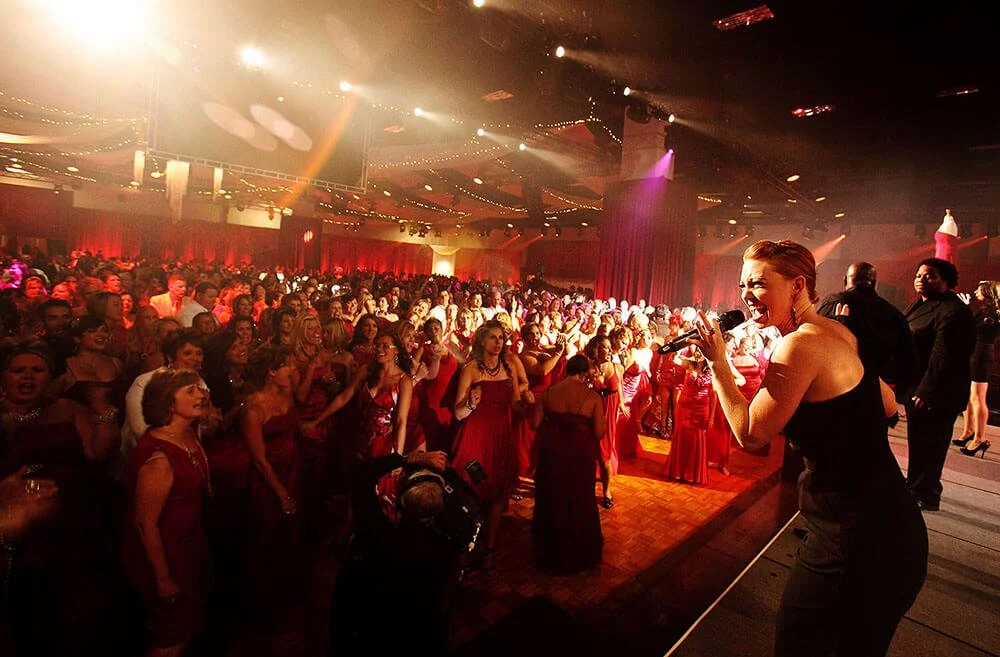 Large crowd of people dancing and enjoying a concert or party under colorful stage lighting.