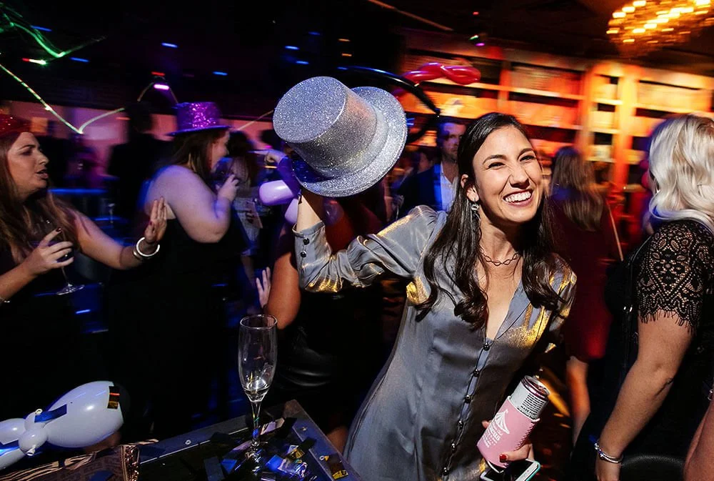 Woman celebrating and holding a sparkling hat in the air, surrounded by partygoers.