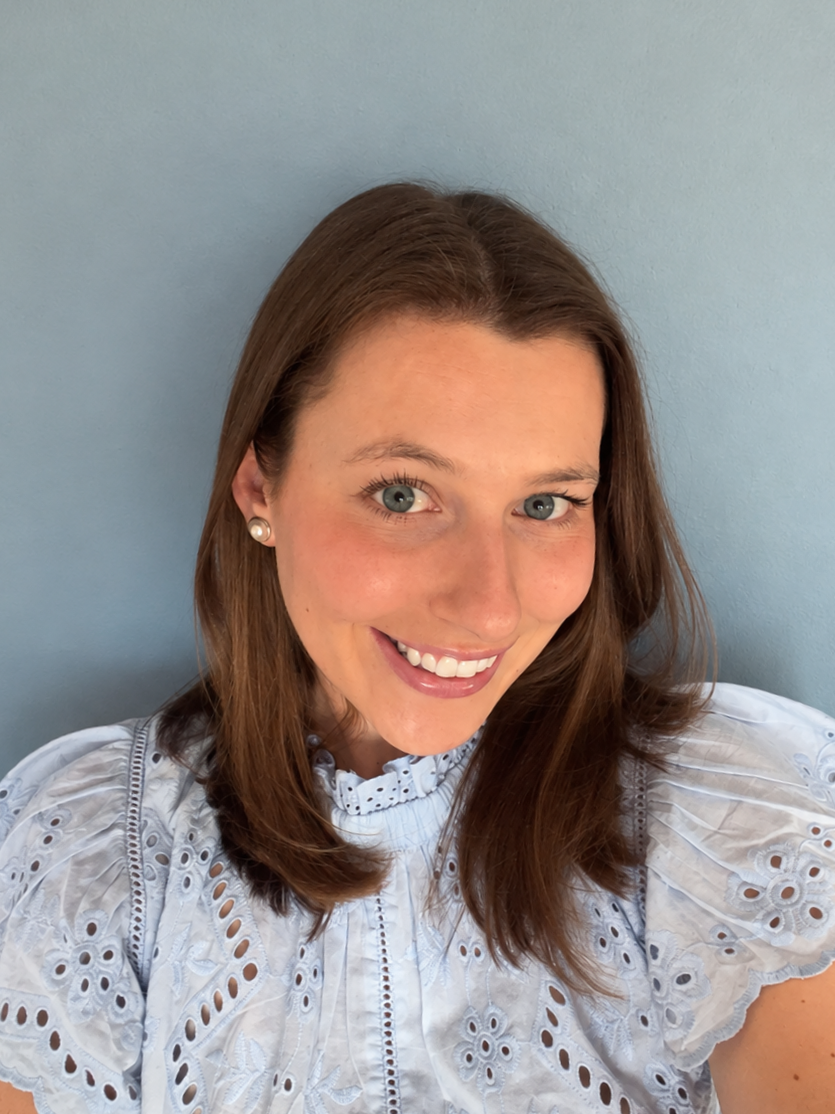 A woman with brown hair, blue eyes, and pearl earrings smiling in front of a blue wall.