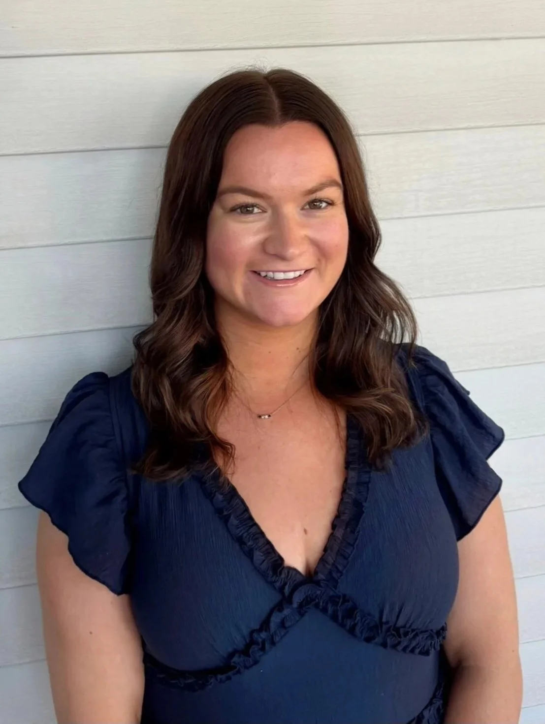 A woman with shoulder-length brown hair smiling, wearing a navy blue top with ruffled sleeves, standing against a light-colored wall with horizontal paneling.