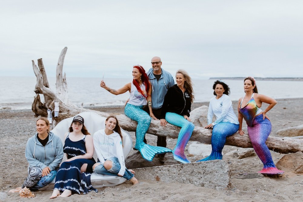 A group of seven women and one man with mermaid tails on a beach posing for a photo near driftwood.