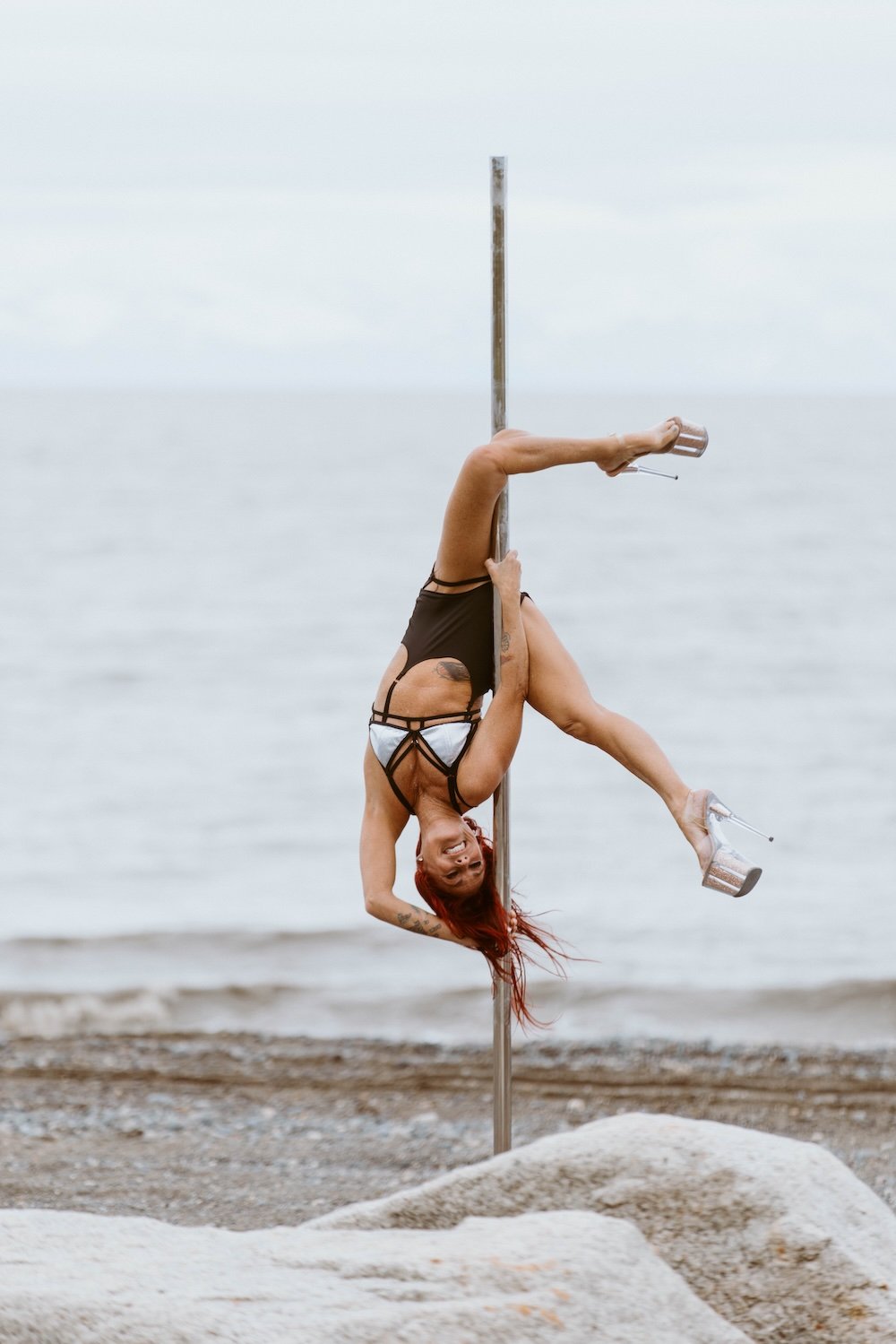 A woman with red hair and tattoos performing a pole dance on a beach. She's upside down, gripping the pole with her legs, wearing high heels, and smiling at the camera. The ocean is in the background.