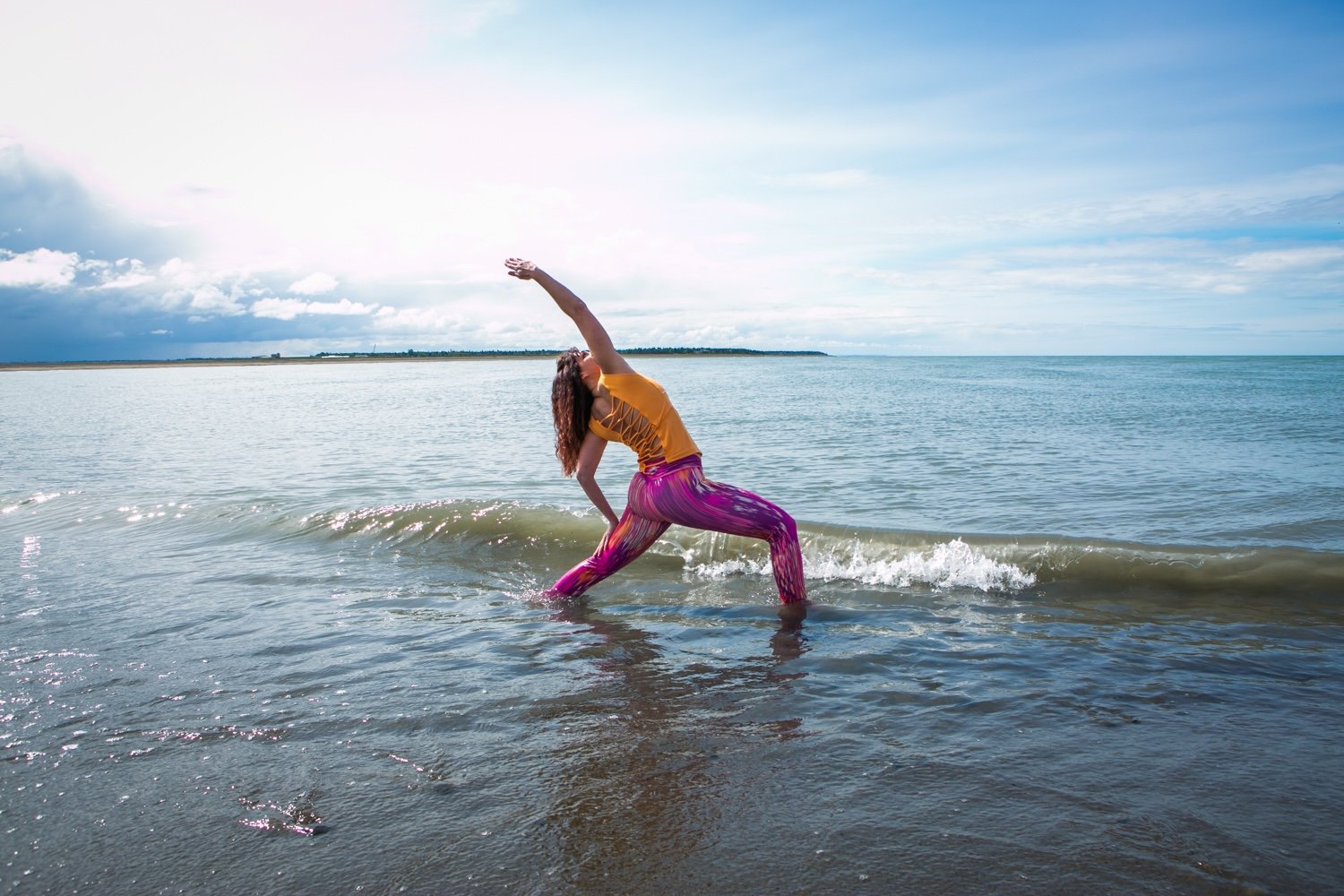 A woman practicing yoga on a beach, standing in shallow water with her left foot forward, right foot back, one arm raised overhead, wearing a yellow top and colorful striped pants, with the ocean and cloudy sky in the background.